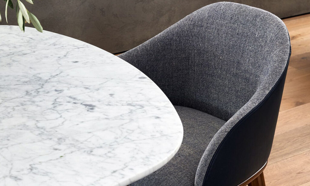A modern dining setup featuring a round white marble tabletop with subtle grey veining, paired with a dark grey upholstered chair with wooden legs. The chair is positioned next to the table on a wooden floor.