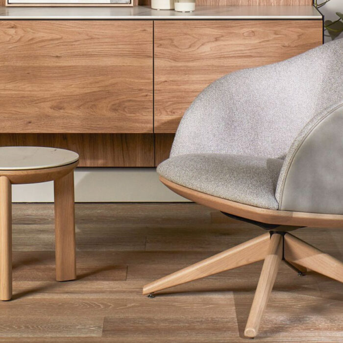 A cozy, modern living space featuring a gray, cushioned swivel chair with wooden legs and a round wooden side table. The backdrop includes a wooden cabinet with clean lines, and the floor is a wooden surface, creating a warm and inviting ambiance.