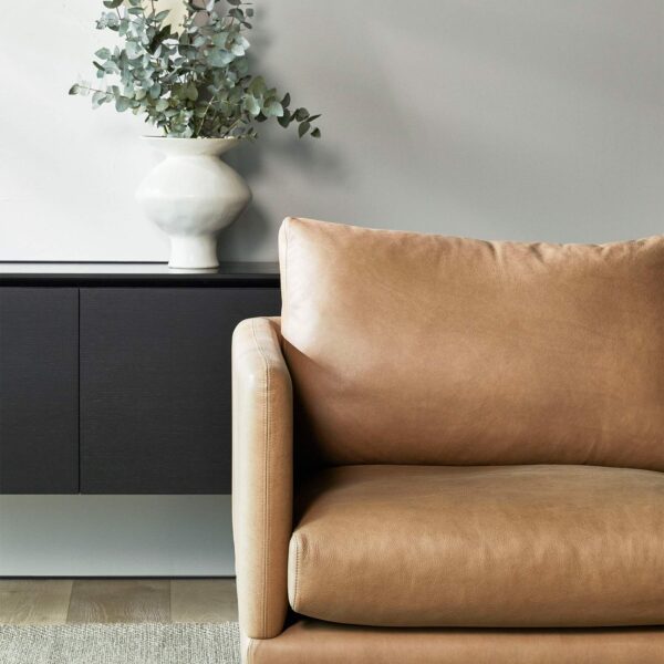 A minimalist living room featuring a tan leather armchair positioned on a beige rug. Behind the chair is a dark modern console table with a white ceramic vase holding eucalyptus branches, set against a light gray wall with a piece of abstract art.