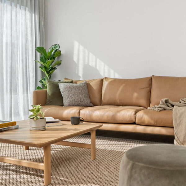 A cozy living room features a light brown leather sofa adorned with throw pillows and a blanket. In front of the sofa is a wooden coffee table with books and a small plant. A grey ottoman sits nearby. The room is brightened by natural light from large windows with sheer curtains.