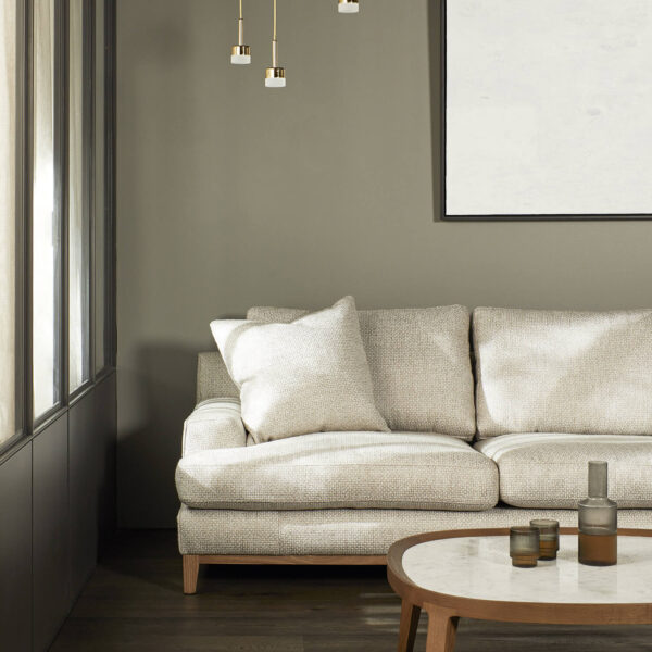 A minimalist living room with a beige sofa, a white and wooden coffee table, and a blank white framed artwork on a gray wall. Several hanging pendant lights with gold accents are above the coffee table. The room features a large window with sheer curtains.