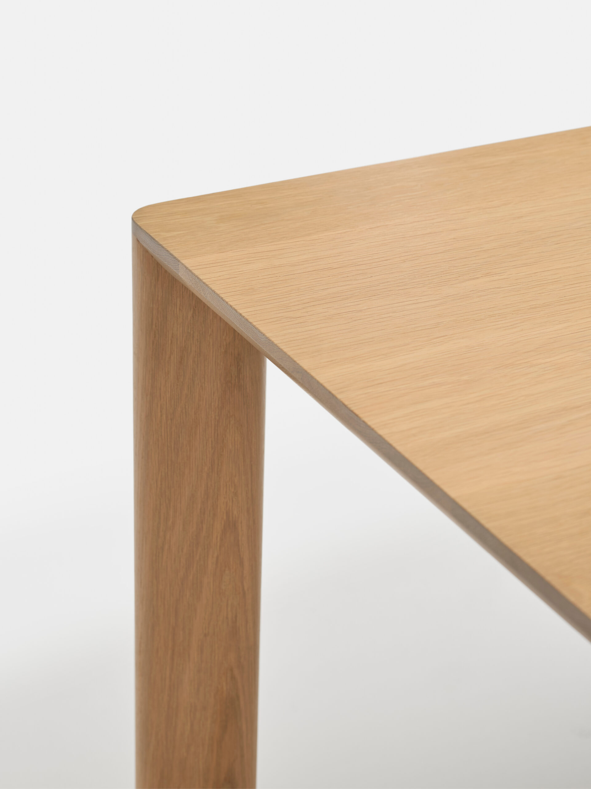 Close-up view of the corner of a wooden table with a smooth, light brown finish, showing the tabletop and one leg against a plain white background.