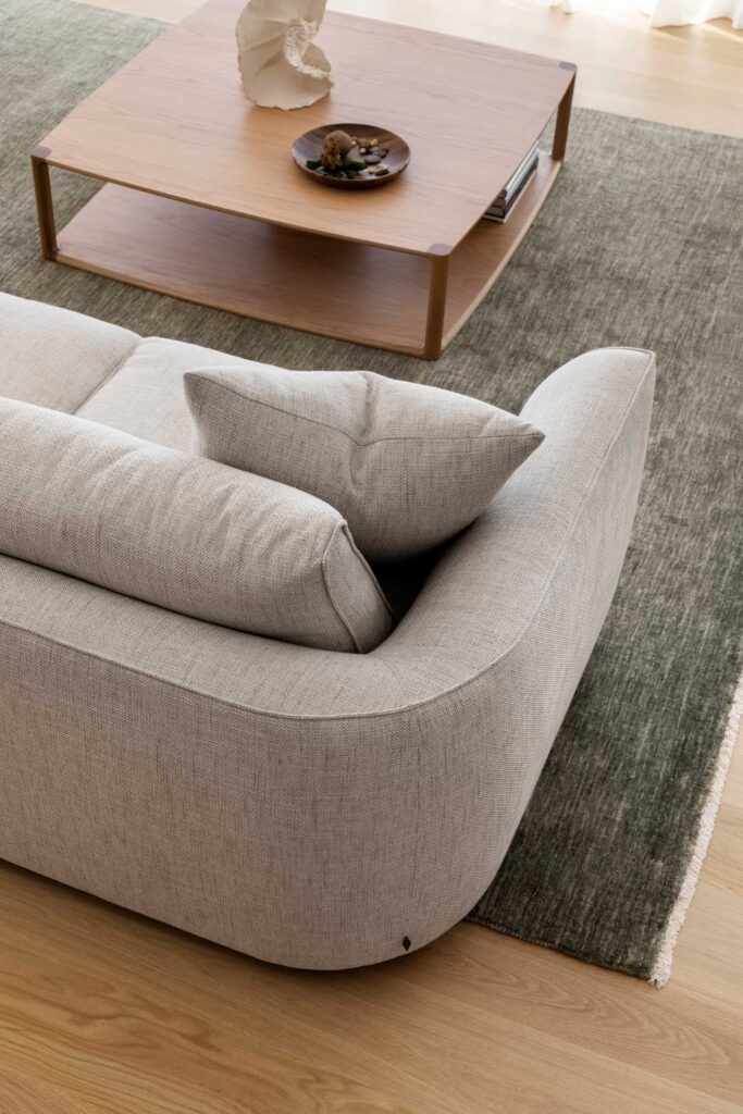 A modern living room with a light gray textured sofa, featuring a square cushion. In front, there's a light wooden coffee table with a decorative bowl and an ornament, resting on a large, greenish-gray area rug that covers a wooden floor.