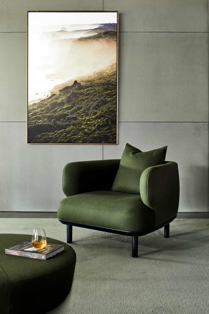 A modern living room features a green armchair with a matching ottoman. A glass of whiskey rests on the ottoman. The walls are light gray, and a large framed photo depicting a misty coastal landscape hangs above the armchair. The room exudes a minimalist aesthetic.