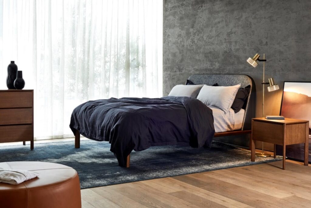 A modern bedroom with a bed dressed in dark bedding, against a textured gray wall. Natural light pours through sheer curtains. The room features a wooden nightstand with a lamp, a dresser with decorative items, a large rug, and a circular ottoman.