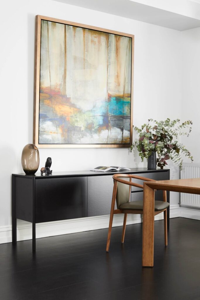 A modern room features a black console table against a white wall, decorated with a large abstract painting in a wooden frame. The table holds a vase, a small sculpture, and an open book. A wooden dining table with a single chair stands on a dark floor. A plant is nearby.