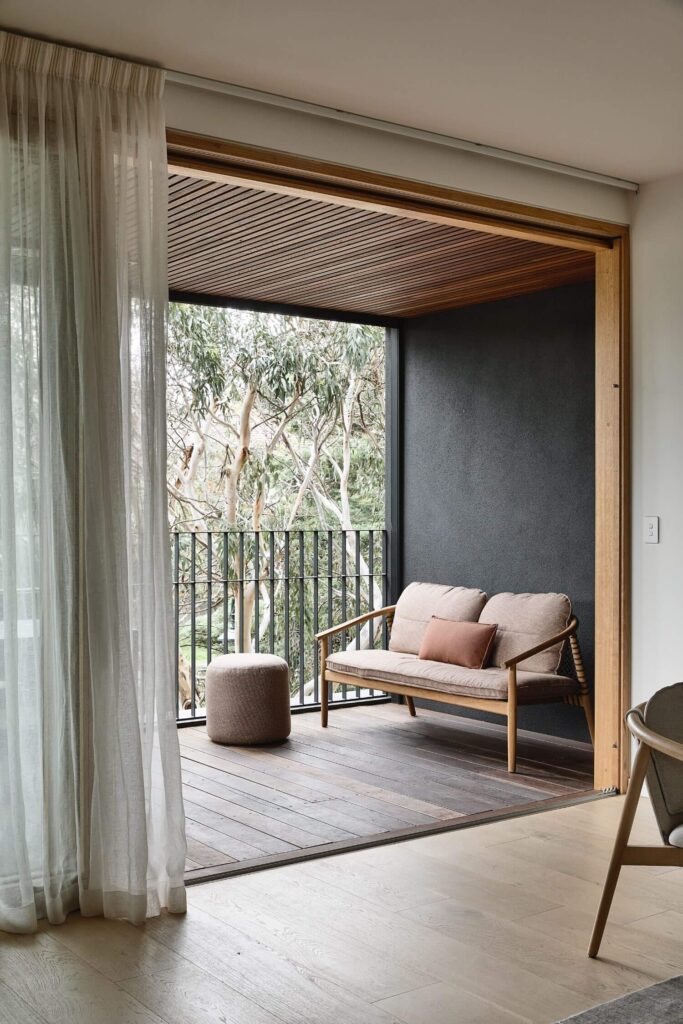 A cozy outdoor balcony features a wooden bench with cushions and a small round ottoman. Light curtains frame the view of tall trees through the railing, creating a serene and inviting atmosphere. The space blends seamlessly with a light wooden indoor floor.