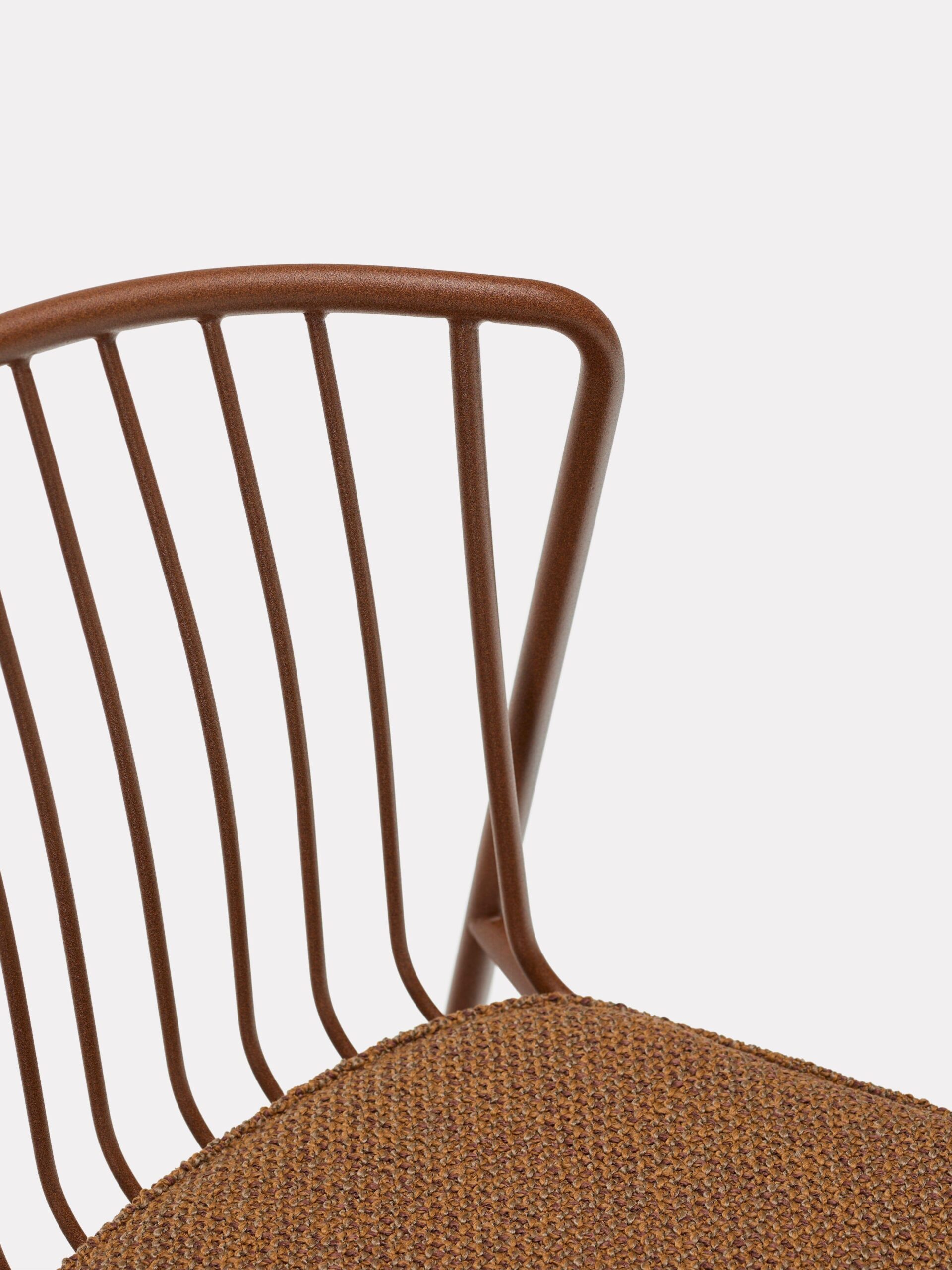 Close-up of a modern chair with a brown metal backrest featuring vertical bars and a textured, brown upholstered seat. The background is plain white.