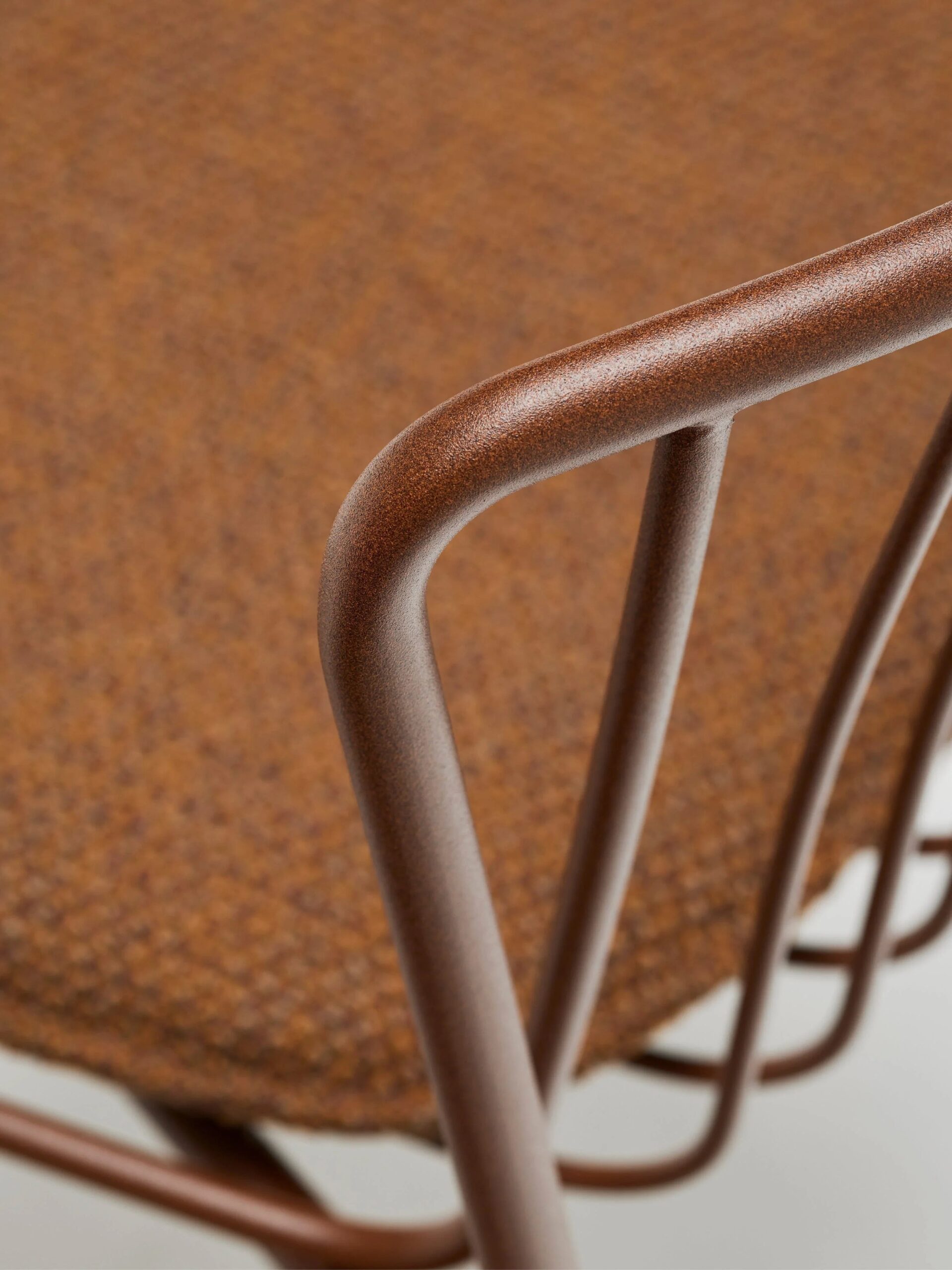 Close-up of a brown metal chair frame and textured brown fabric seat, showing the smooth curves and minimalist design of the chair’s armrest and support structure.