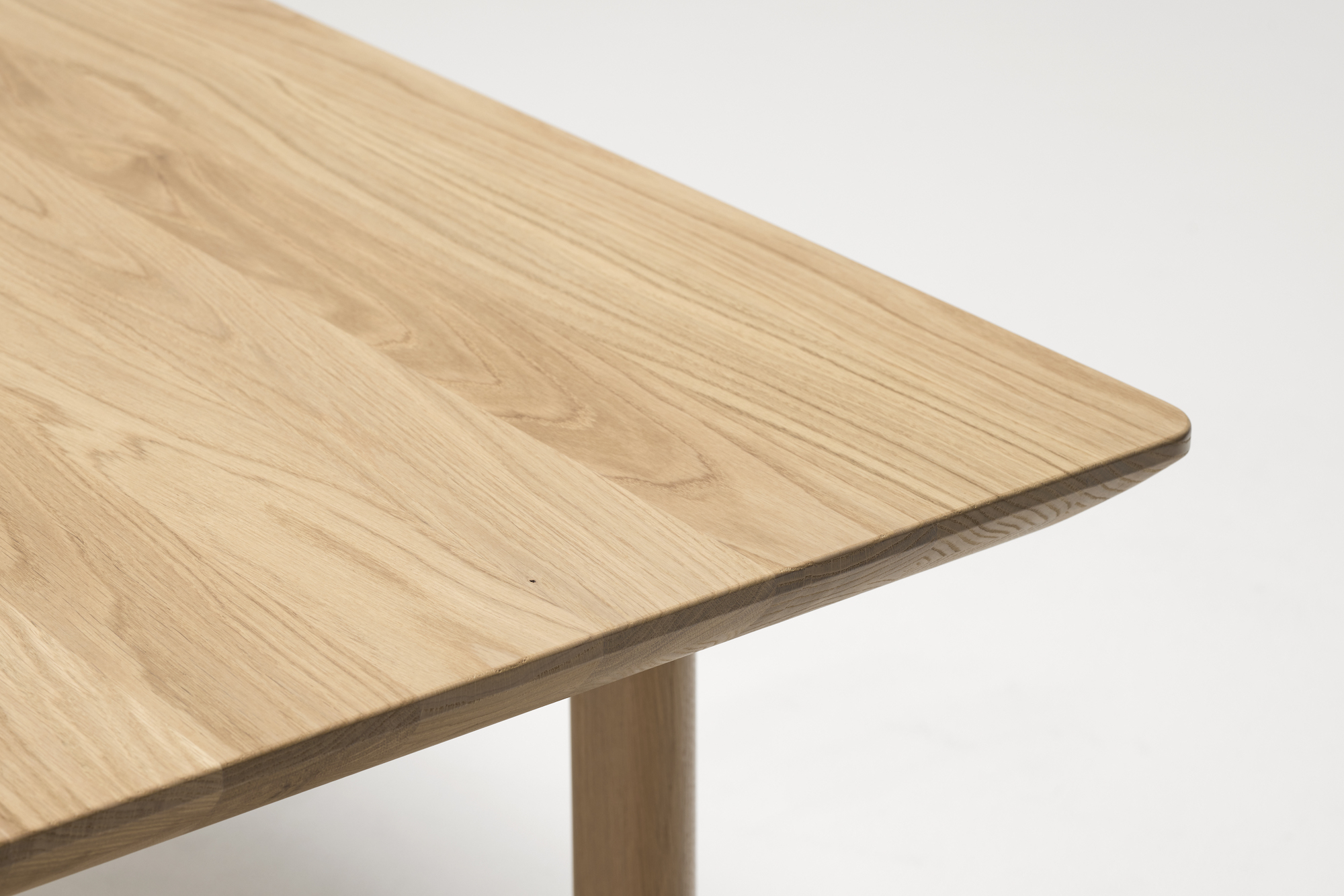 A close-up view of the corner of a wooden table made from light-colored timber furniture. The table's surface has visible wood grain patterns, and the corner has smooth, curved edges. The background is plain white.