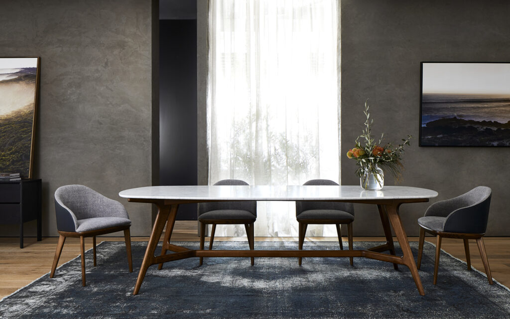 Modern dining room featuring a marble-top oval table with wooden legs, surrounded by five gray upholstered chairs. A bouquet of flowers in a glass vase sits on the table. Large window with sheer curtains in the background, and two framed landscape photos adorn the walls, highlighting elegant marble tables.