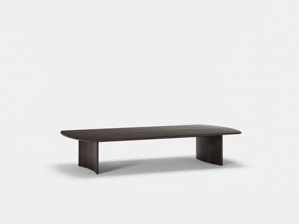 A minimalist, rectangular dark wood coffee table with a smooth surface and slightly rounded edges is centered against a plain white background. The table has two wide, flat legs at each end, creating a sleek and modern appearance.
