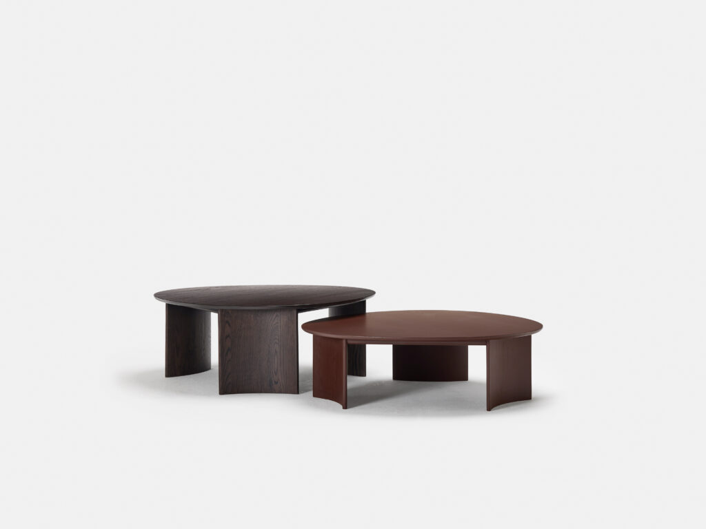 Two sleek, round, modern coffee tables with simple, curved legs are placed beside each other against a plain white background. One table is dark brown with a wood grain texture, and the other is a smooth, lighter reddish-brown.