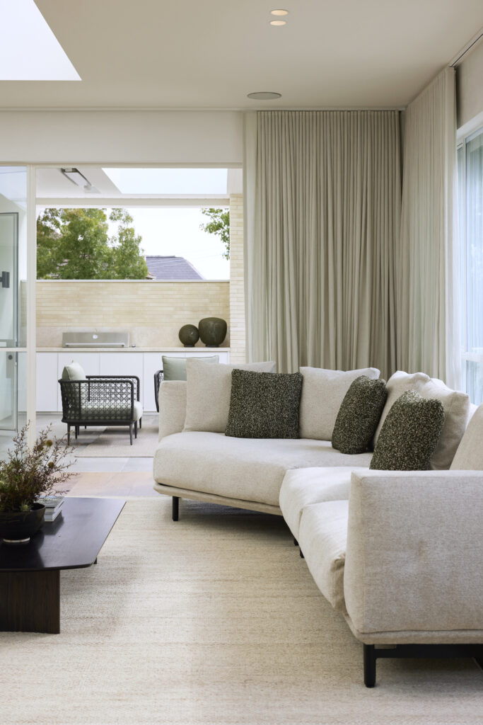 A modern living room with a light gray Feathertop sectional sofa adorned with patterned pillows. The room has a sleek coffee table and soft beige area rug. A sliding glass door opens to a patio with outdoor seating and a built-in grill. Light curtains frame the large windows.