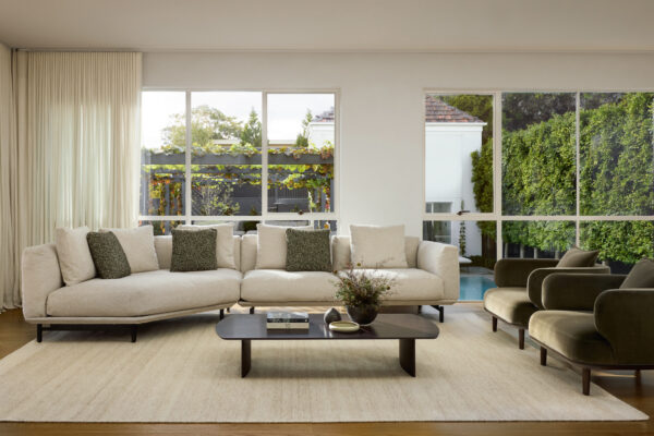 A modern living room featuring the Feathertop Sofa, a large beige sectional adorned with green and beige cushions, flanked by two green armchairs. A low wooden coffee table with flowers rests on a light rug. Large windows reveal a view of a lush garden and pool outside.