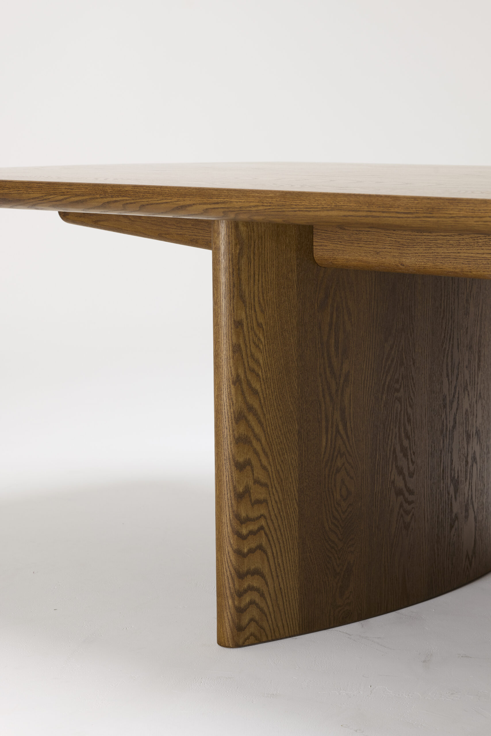 A close-up of a modern wooden table with a smooth surface and a thick, curved leg, showcasing the natural wood grain and clean, minimalist design. The background is plain and white.