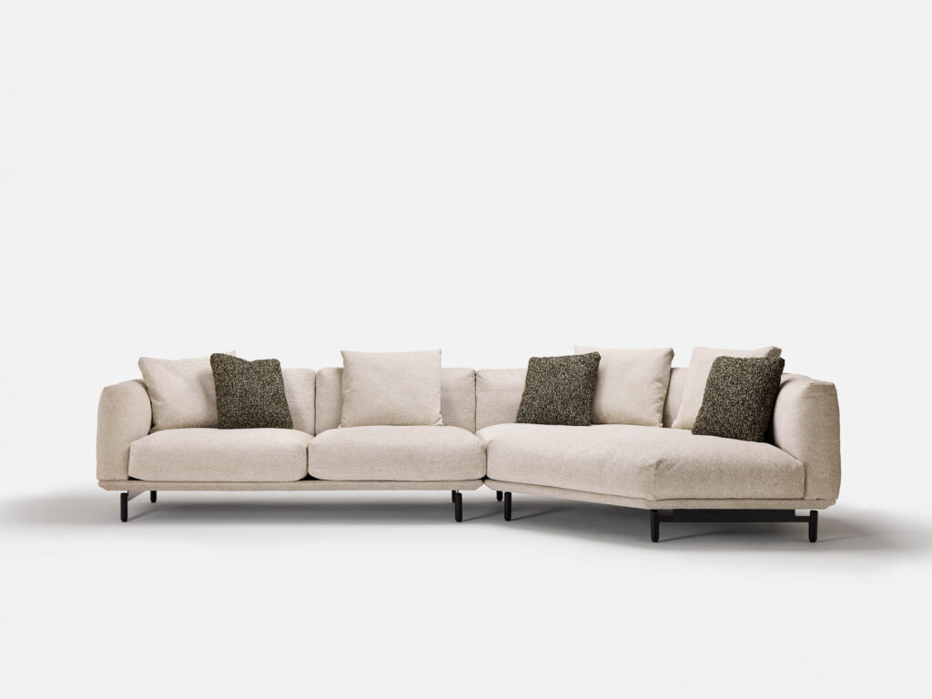 A modern beige Feathertop Sofa with a chaise lounge on the right side. The sofa features four seat cushions, back cushions, and four decorative dark green throw pillows. It has slim, black legs, creating a minimalist design against a plain white background.