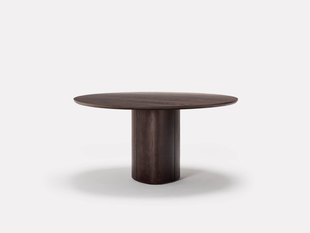 A minimalist round wooden table with a dark brown finish. The tabletop is smooth and supported by a single, cylindrical pedestal base. The background is plain white.