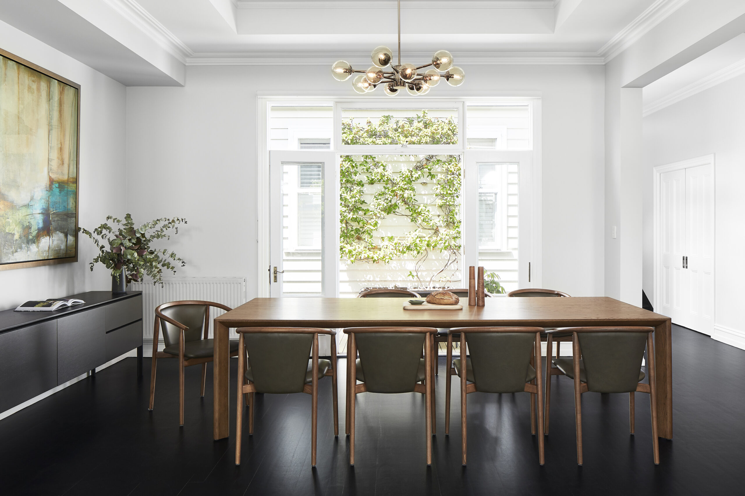 A modern dining room with a wooden table surrounded by eight green cushioned chairs. A contemporary chandelier hangs above the table, and a large window with glass doors reveals a lush outdoor scene. A sideboard with decor items sits against the white walls.
