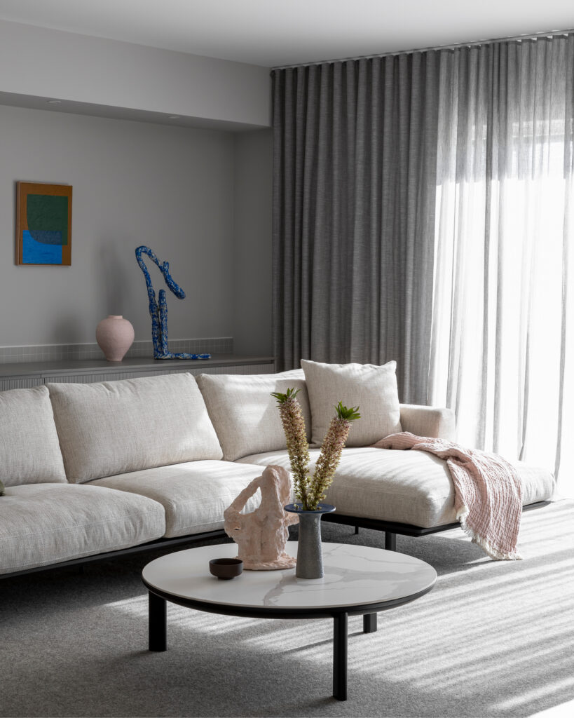 A modern living room with a light gray L-shaped sofa adorned with a pink throw blanket. A round coffee table with vases of decorative plants sits in front. The back wall has artwork, a blue sculpture, and a pink vase. Floor-to-ceiling gray curtains let in soft light.