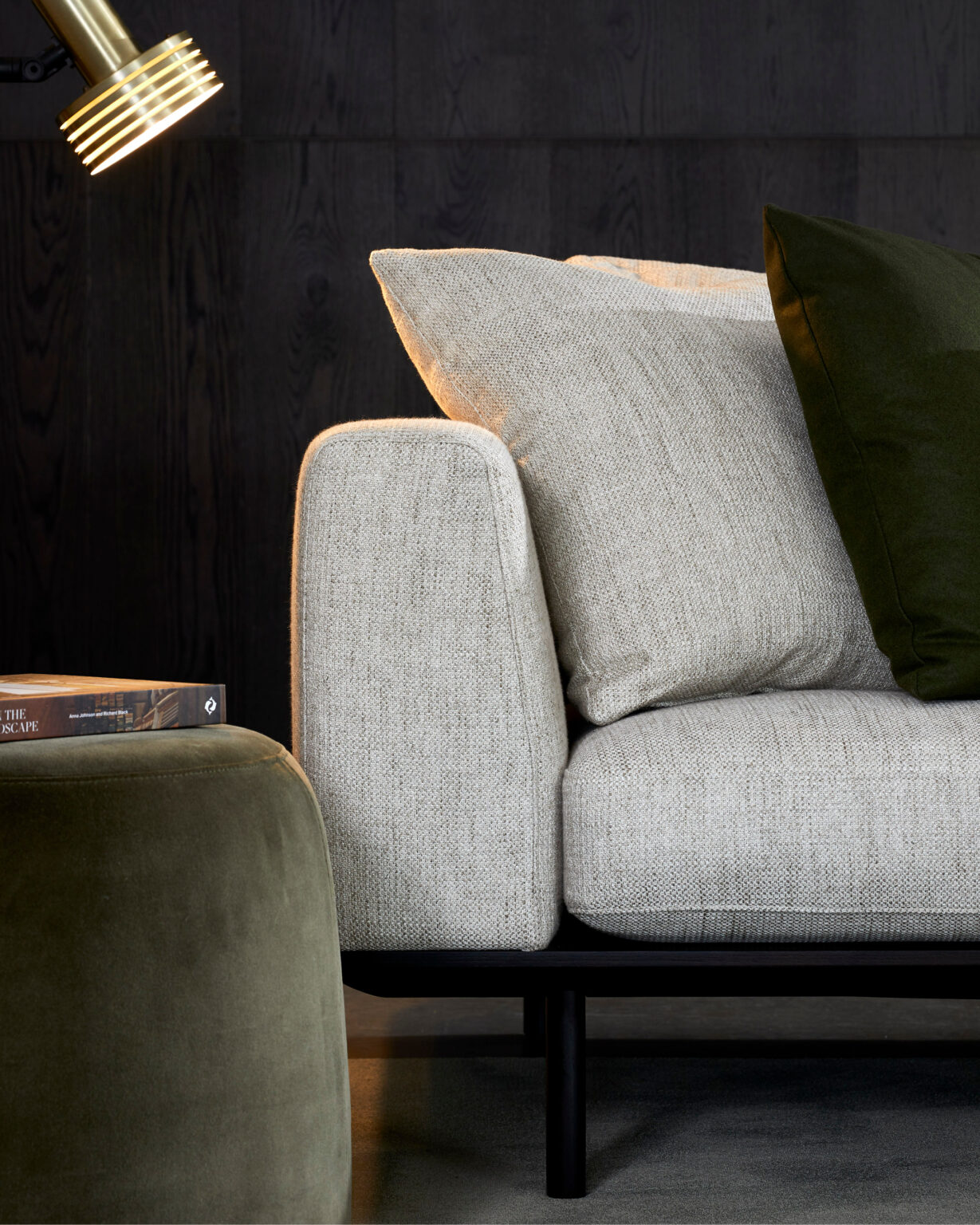 A cozy modern living room scene featuring a light gray sofa with square armrests, adorned with light gray and dark green pillows. Nearby is an olive green ottoman and a stack of books. A brass floor lamp emits a warm light, illuminating the scene.