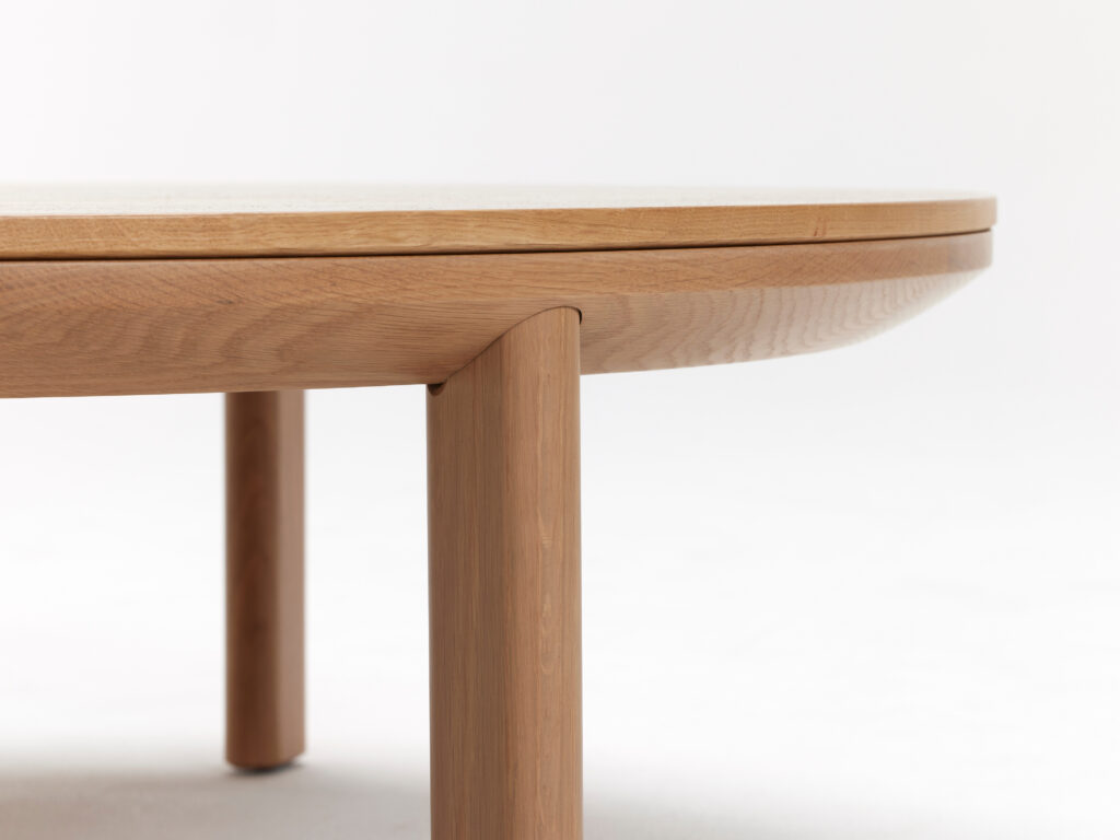 Close-up of a wooden tabletop and its supporting legs. The image focuses on the natural wood grain and the smooth, rounded edge of the table, highlighting the craftsmanship and light wood finish. The background is plain white.