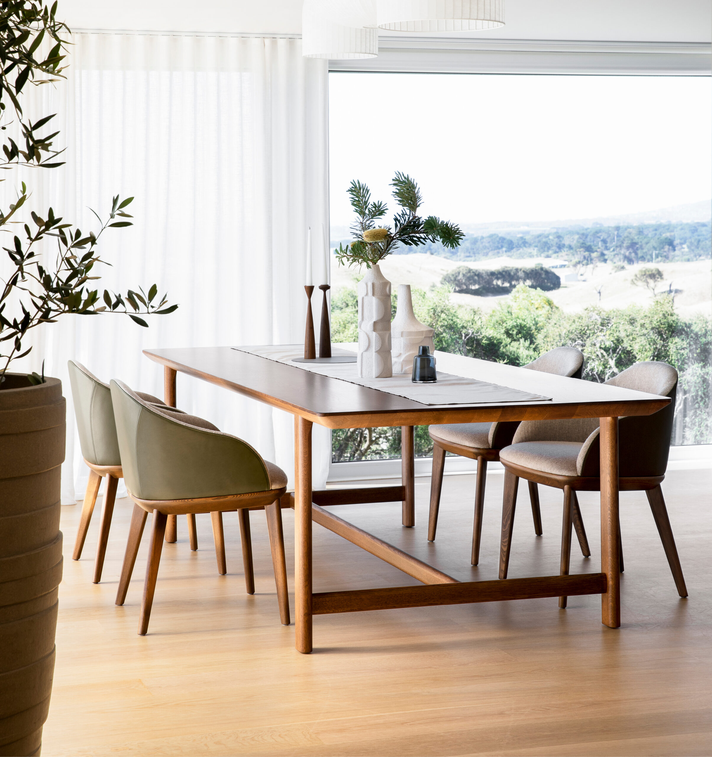 A modern dining area with a wooden table set for six, featuring light olive-green cushioned chairs. A minimalist table runner holds a white vase with greenery, a blue vase, and two tall wooden candlesticks. The background showcases a large window with a scenic view.