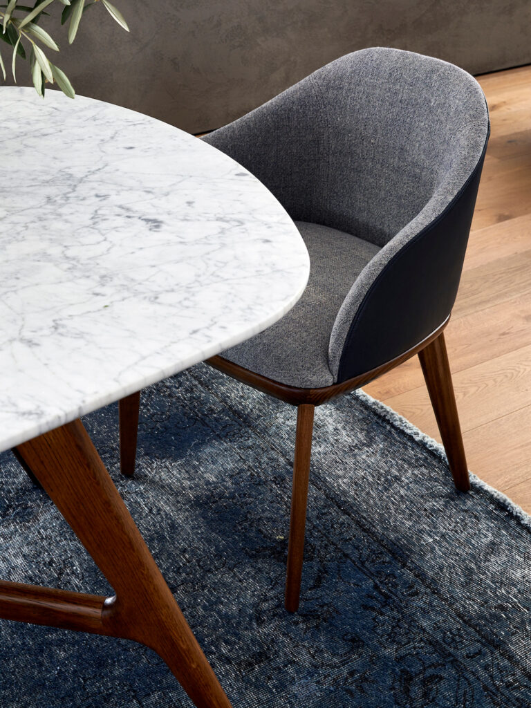A modern dining chair with a gray upholstered seat and wooden legs sits next to a round white marble dining table on a dark blue rug, atop a wooden floor.