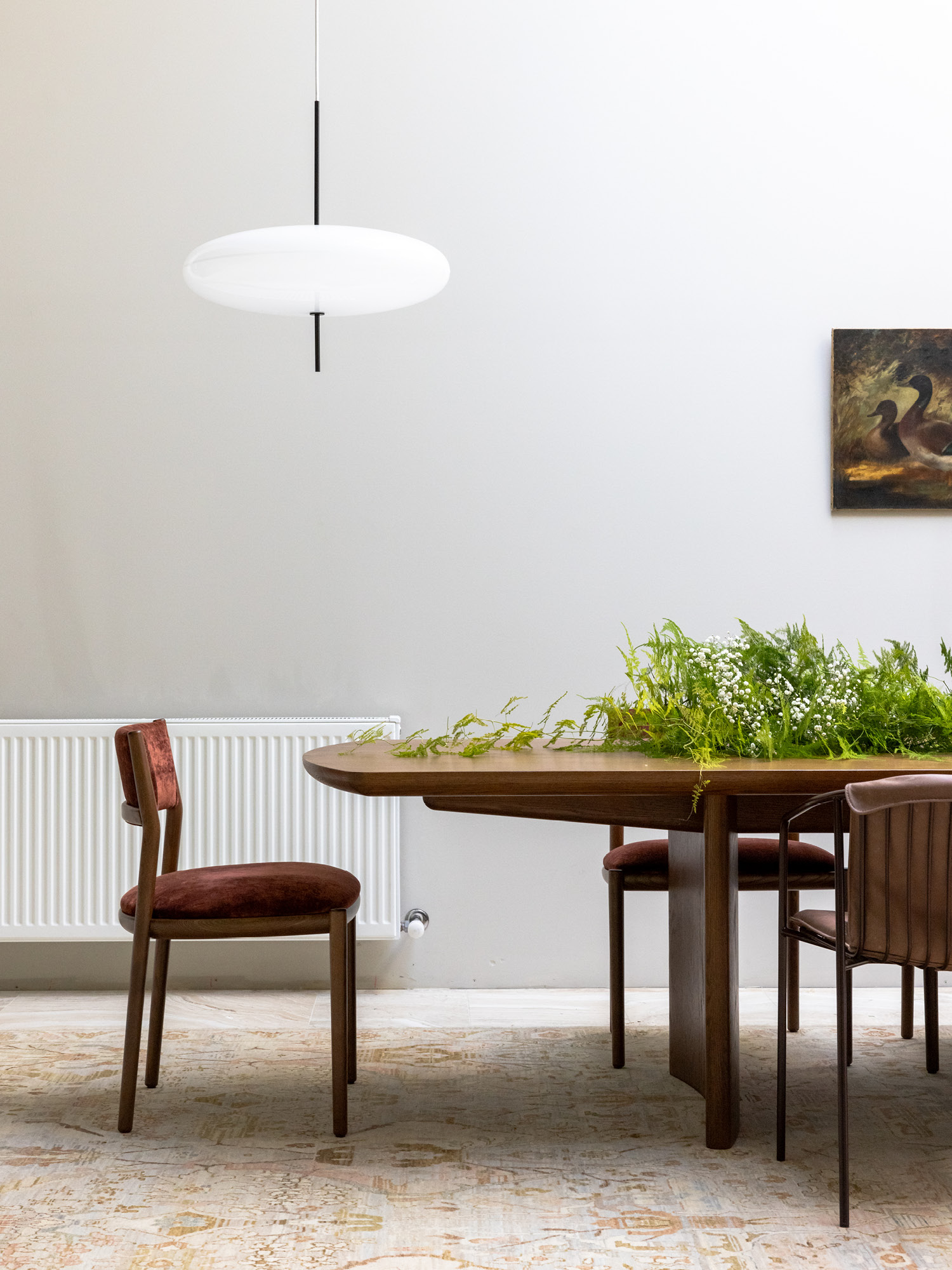 A modern dining room with a dark wooden table, brown velvet chairs, green plants arranged on the table, a white pendant light above, a radiator on the wall, and a painting of birds hanging in the background.