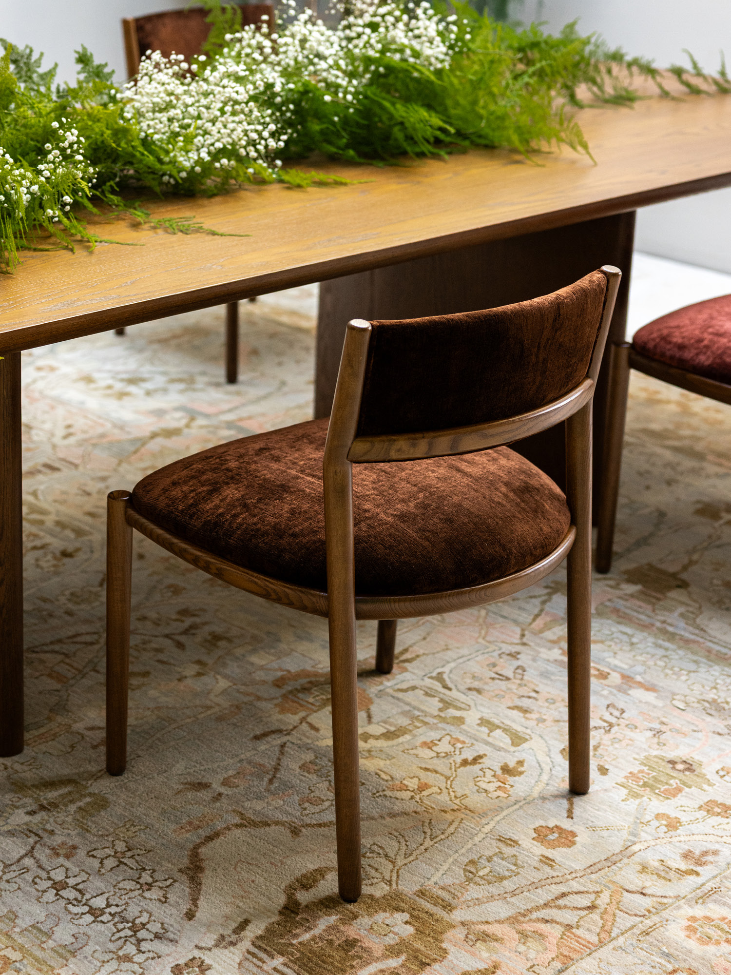 A wooden dining chair with a brown velvet seat is placed at a wooden table decorated with green ferns and white flowers, sitting on a patterned beige rug.