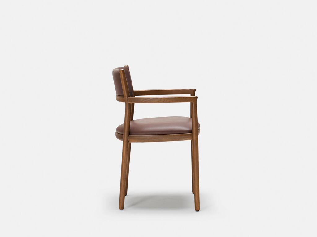 A wooden chair with a brown leather seat and backrest, shown in profile against a plain white background. The chair has armrests and simple, clean lines in its design.
