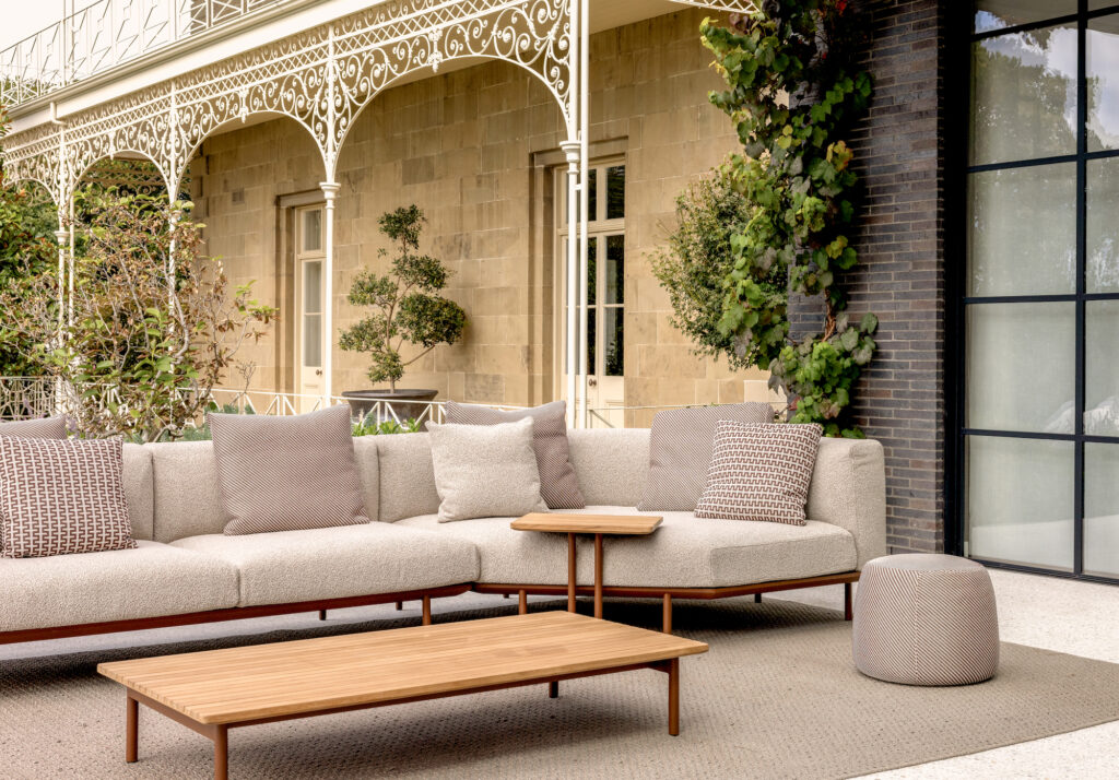An outdoor patio with a beige sectional sofa, patterned and solid cushions, a wooden coffee table, a small side table, and a round ottoman. The area is set against a building with ornate trim, brick walls, and large windows.