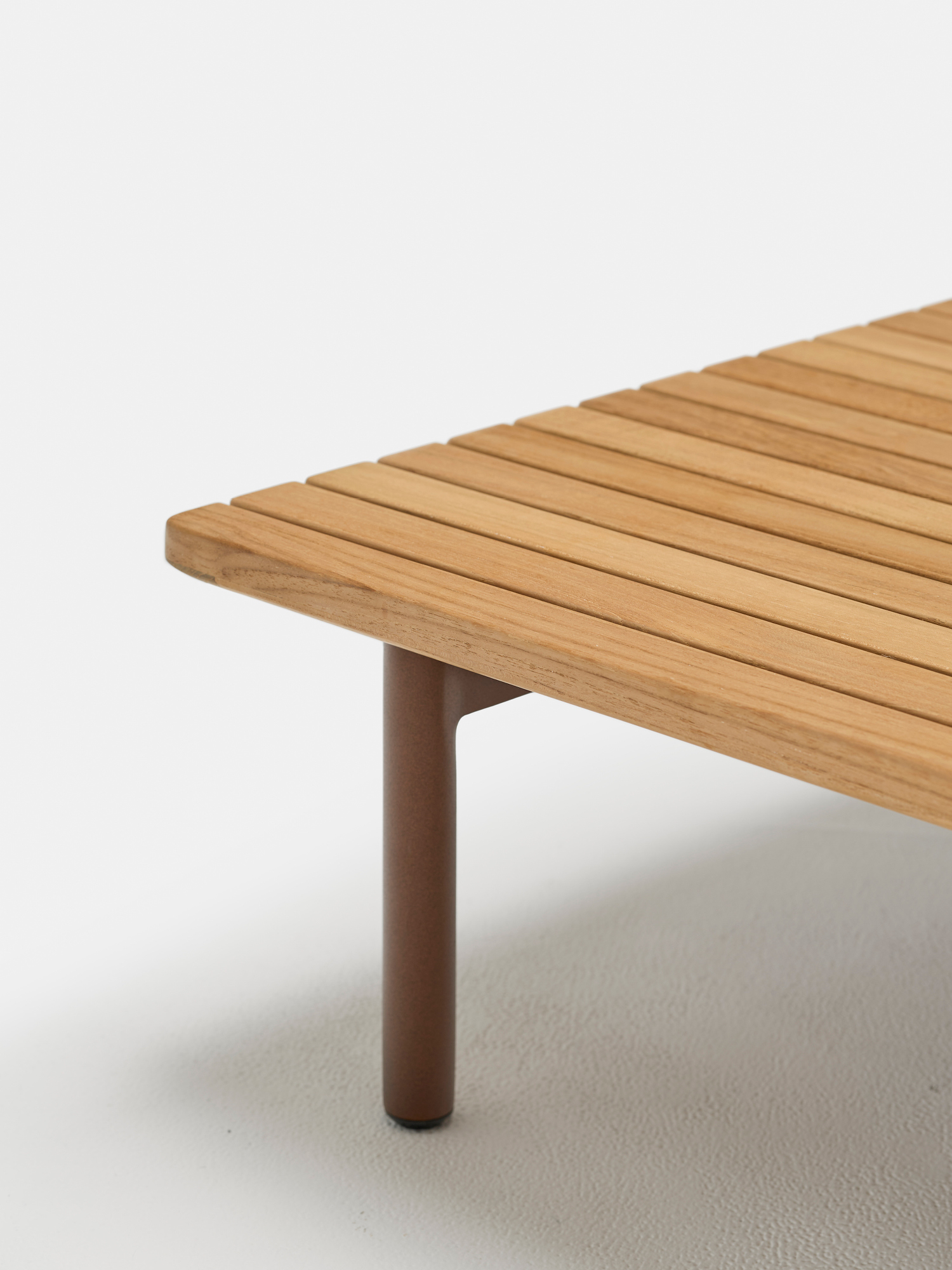 A close-up of the corner of a wooden slatted bench with a rounded brown metal leg, set against a plain white background.