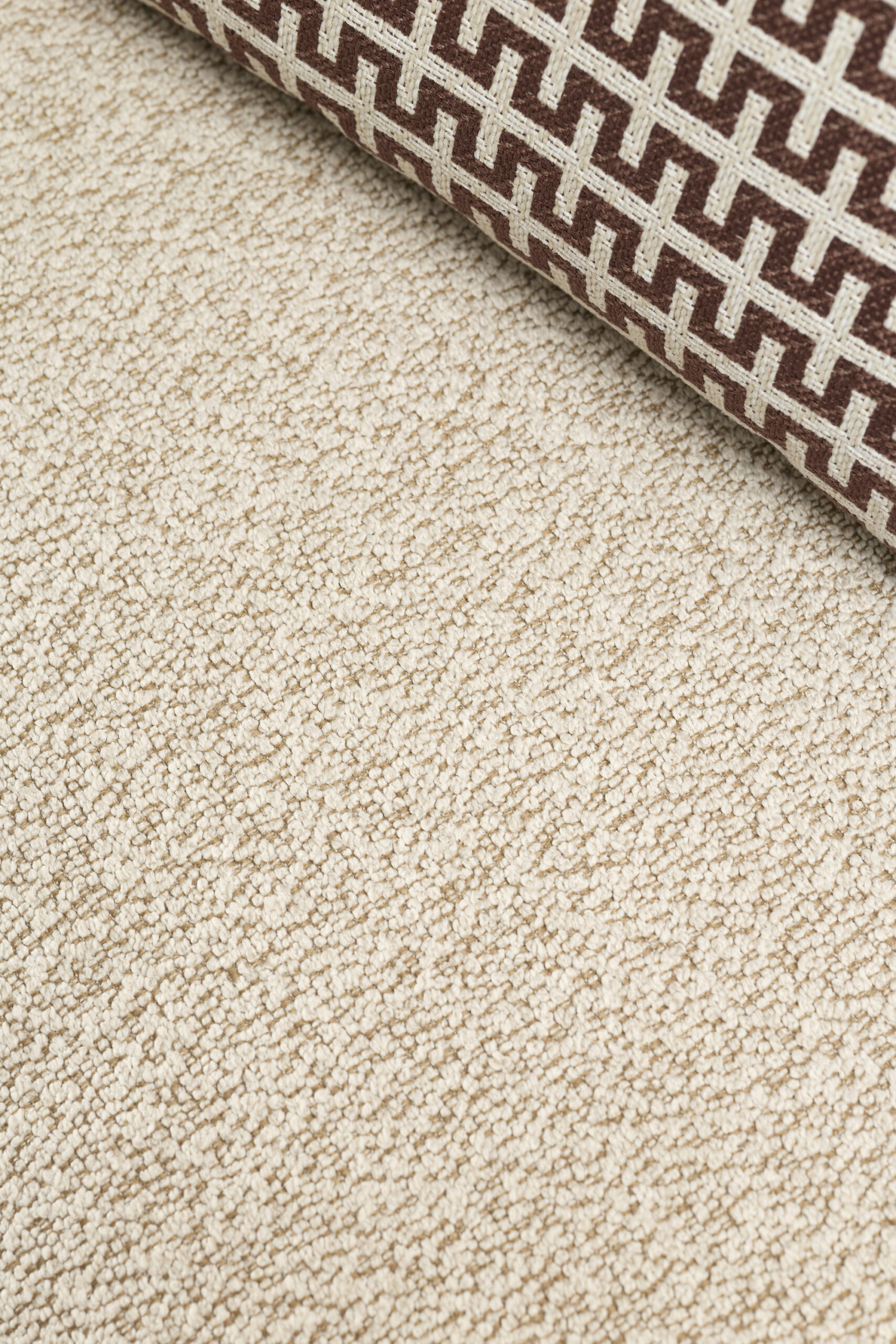 A close-up of a beige textured carpet with a rolled-up section featuring a geometric brown and beige pattern in the corner.