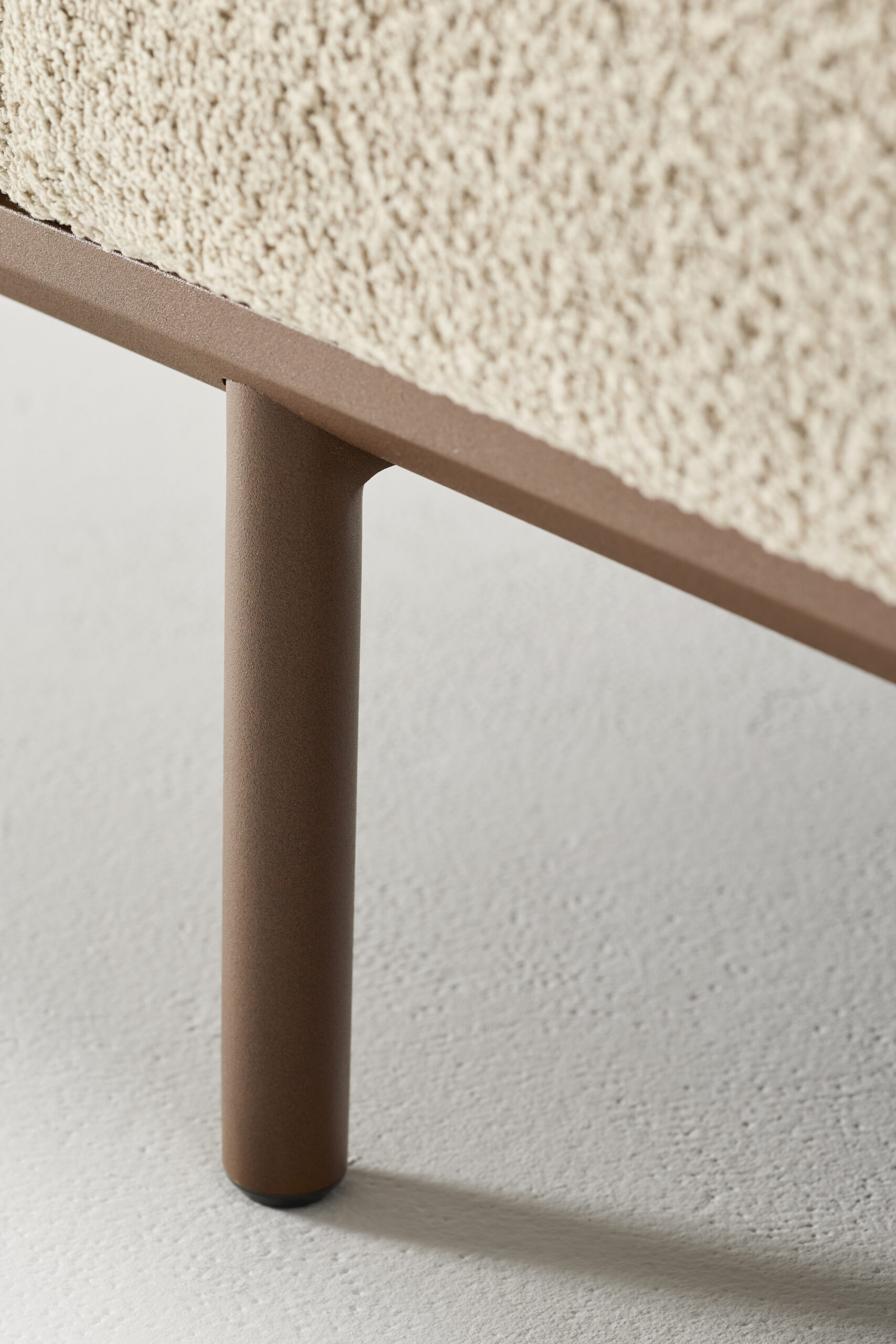 Close-up of a modern furniture leg in matte brown metal, supporting a piece covered in textured beige fabric, standing on a light-colored, smooth floor.
