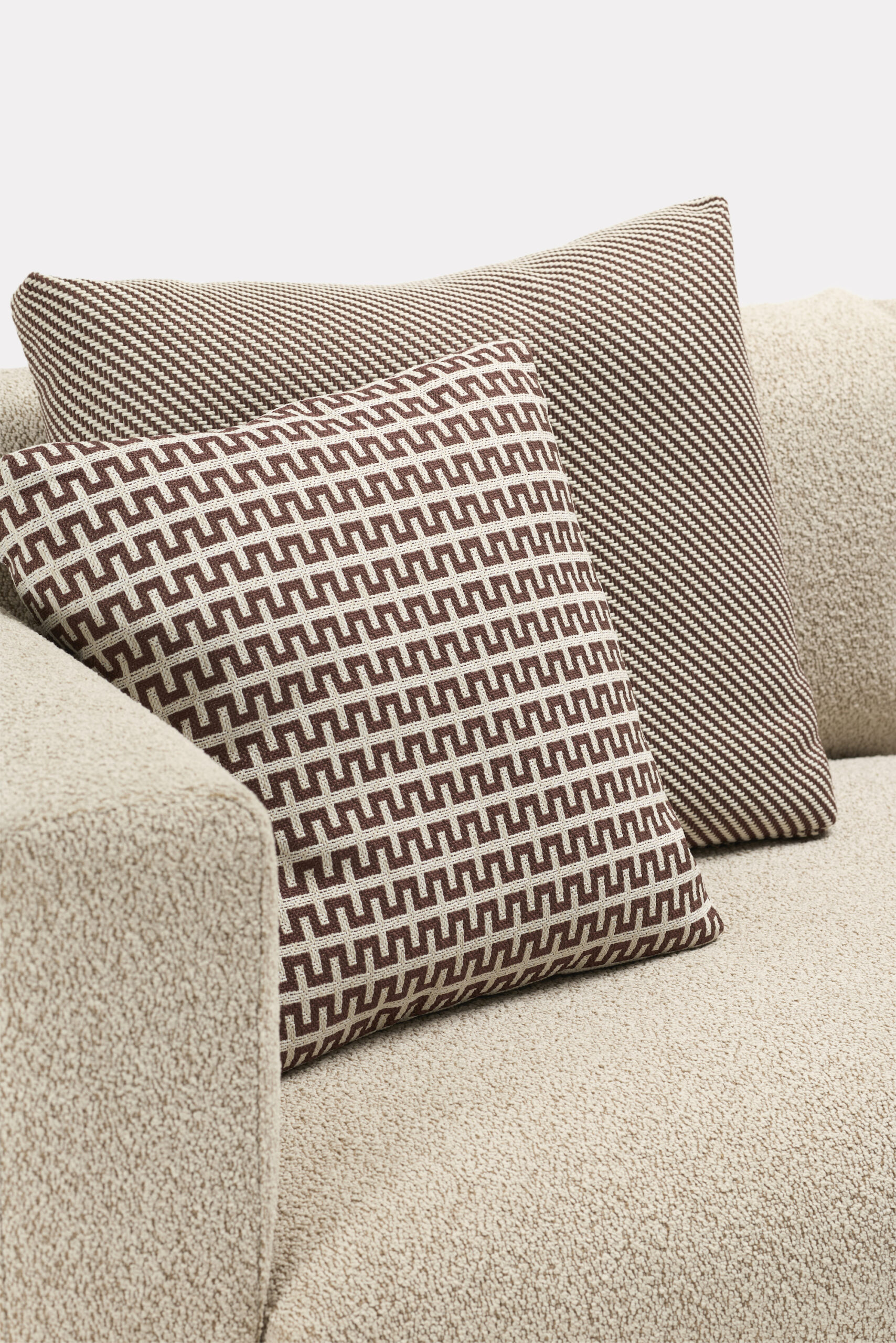 Two decorative cushions with brown and beige geometric patterns rest on the arm of a textured beige sofa against a plain white background.