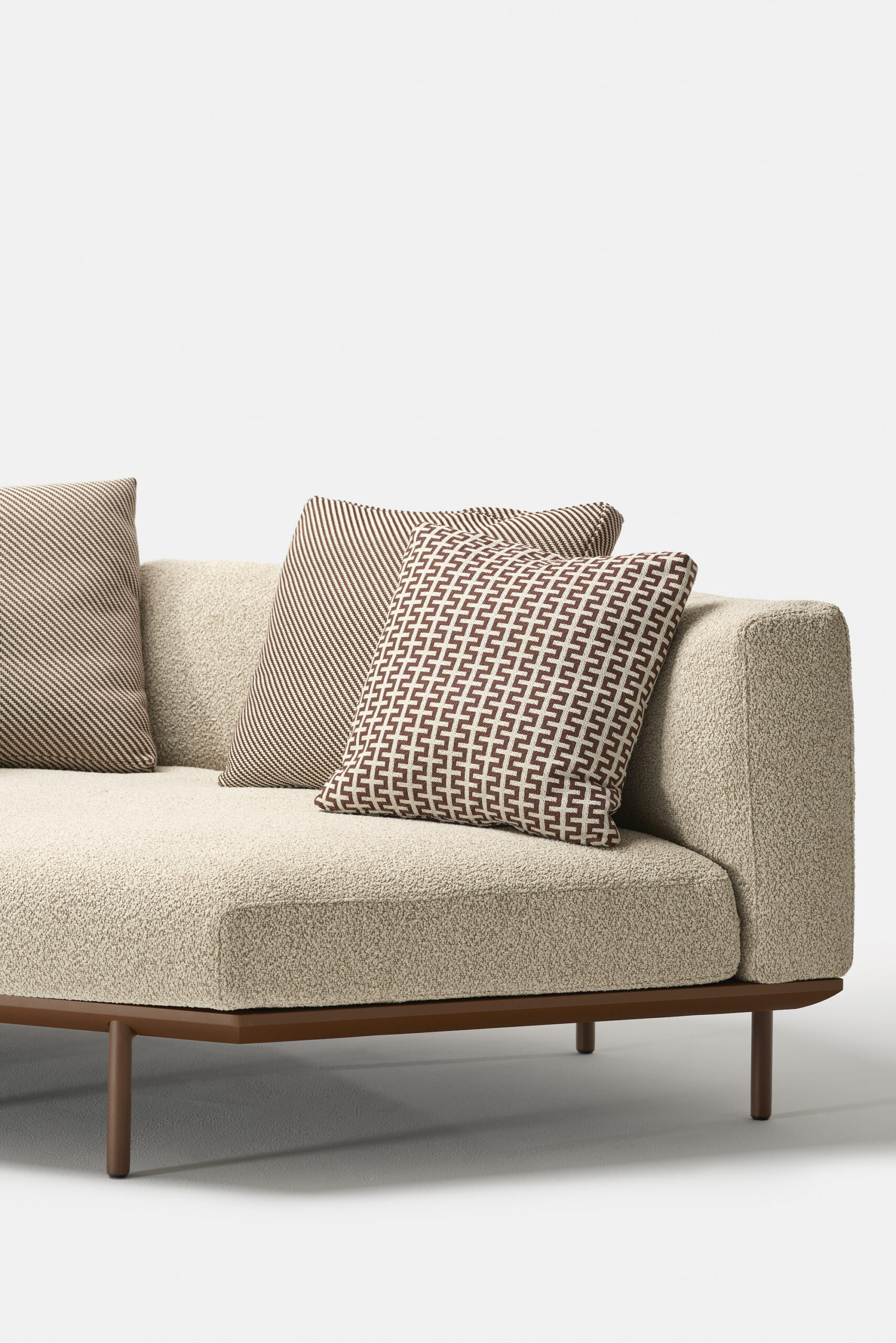 A modern beige sofa with textured upholstery, featuring a wooden base and three decorative pillows in brown and white geometric patterns, set against a plain white background.