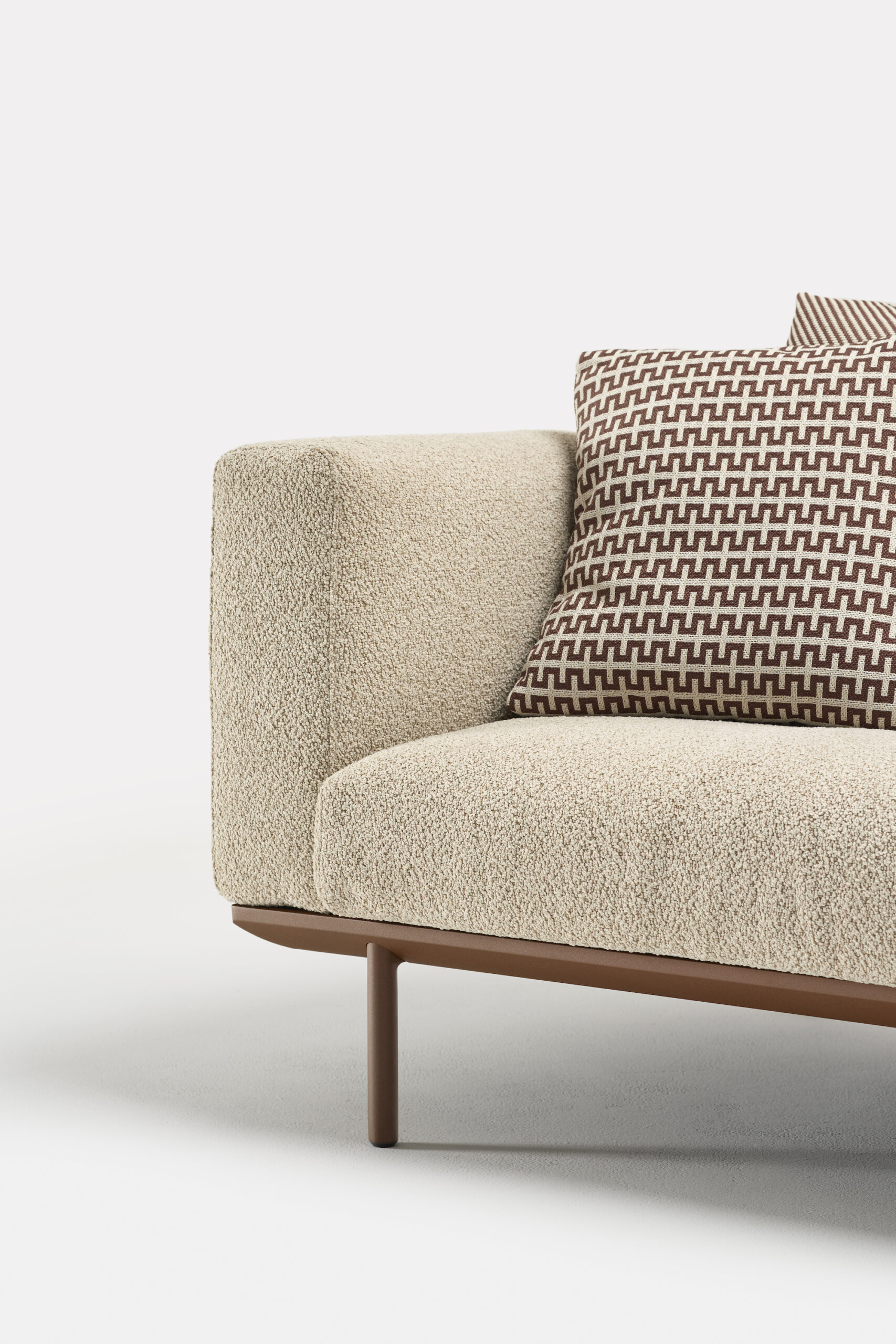 A modern beige fabric sofa with a brown wooden base, featuring two brown-and-white geometric patterned cushions placed on the seat. The image shows a close-up of the sofa's left side against a plain background.