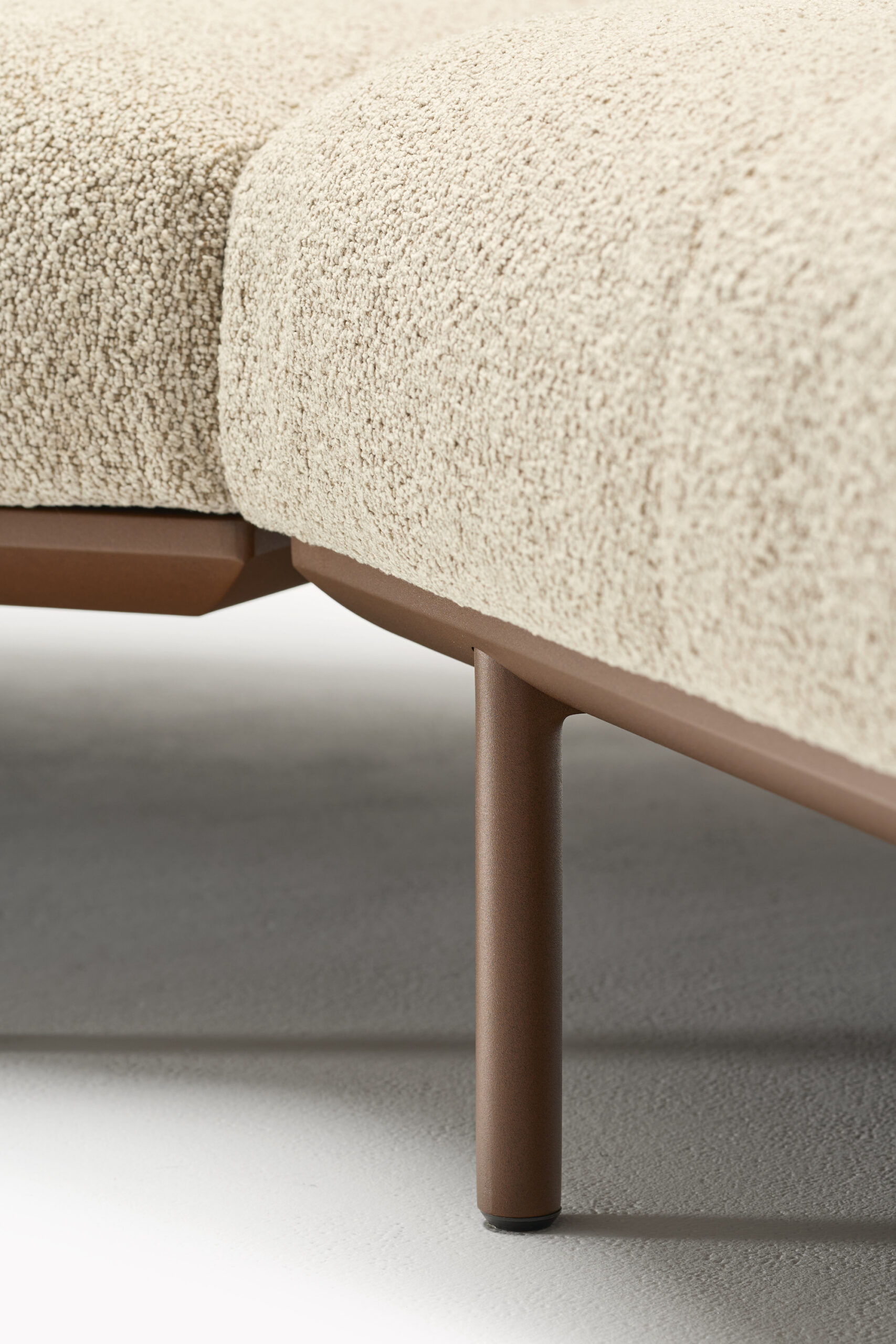 Close-up of a modern beige upholstered sofa with textured fabric, showing two seat cushions and a brown metal leg on a light-colored floor.