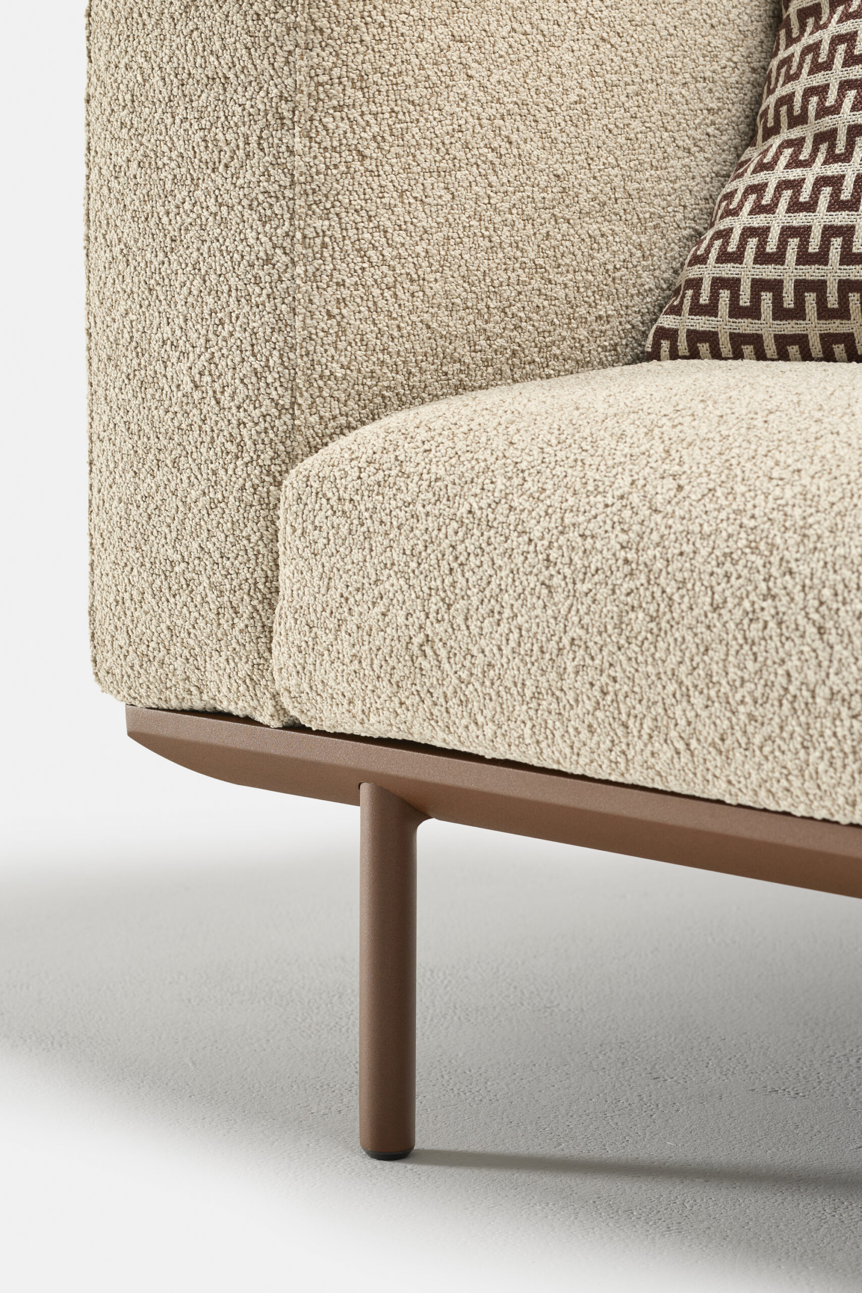 Close-up of a modern beige upholstered sofa with textured fabric, featuring a brown metal leg and a decorative cushion with a geometric pattern in brown and white.