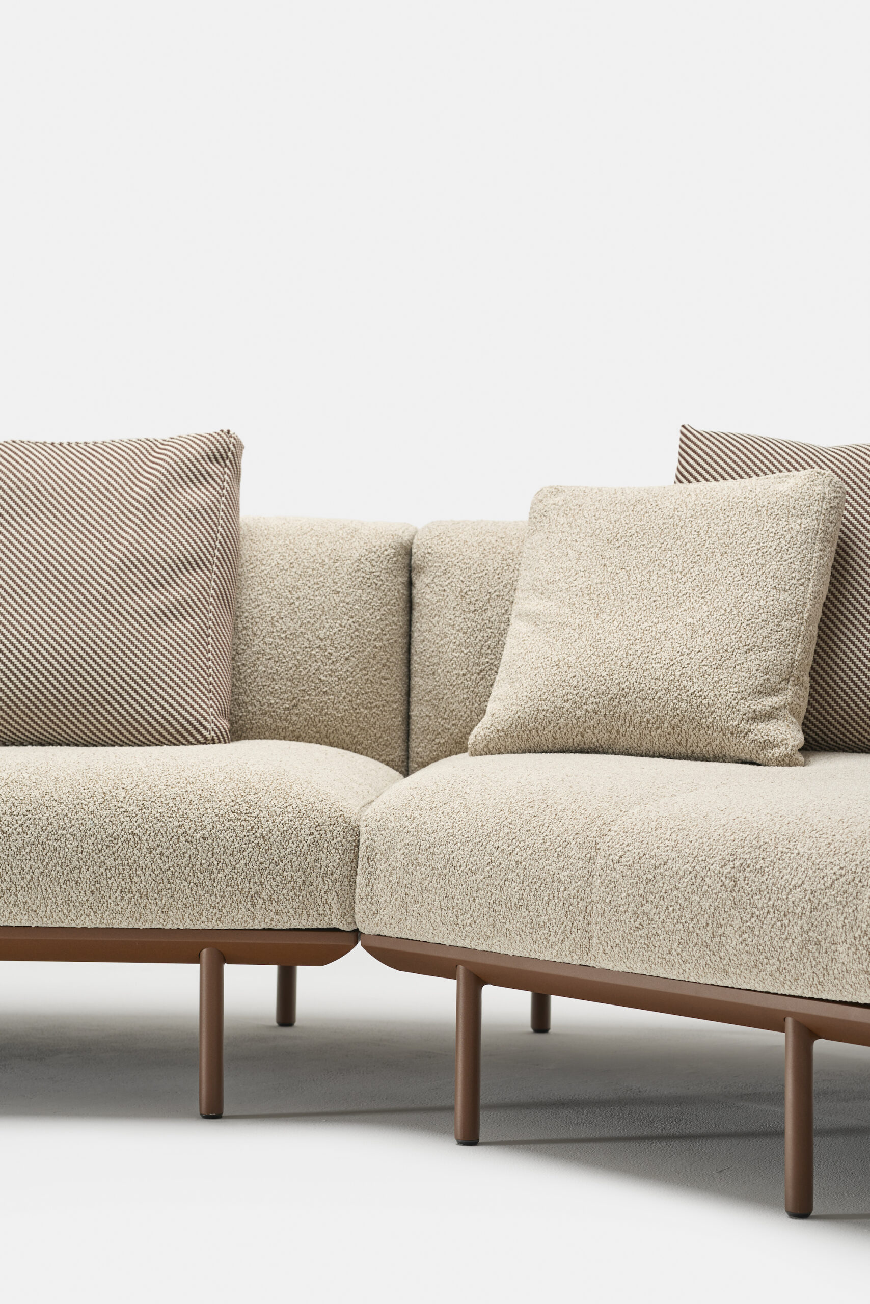 A modern, beige L-shaped sofa with textured upholstery and brown legs, featuring two cushions—one with a solid color and one with a striped pattern—against a plain white background.