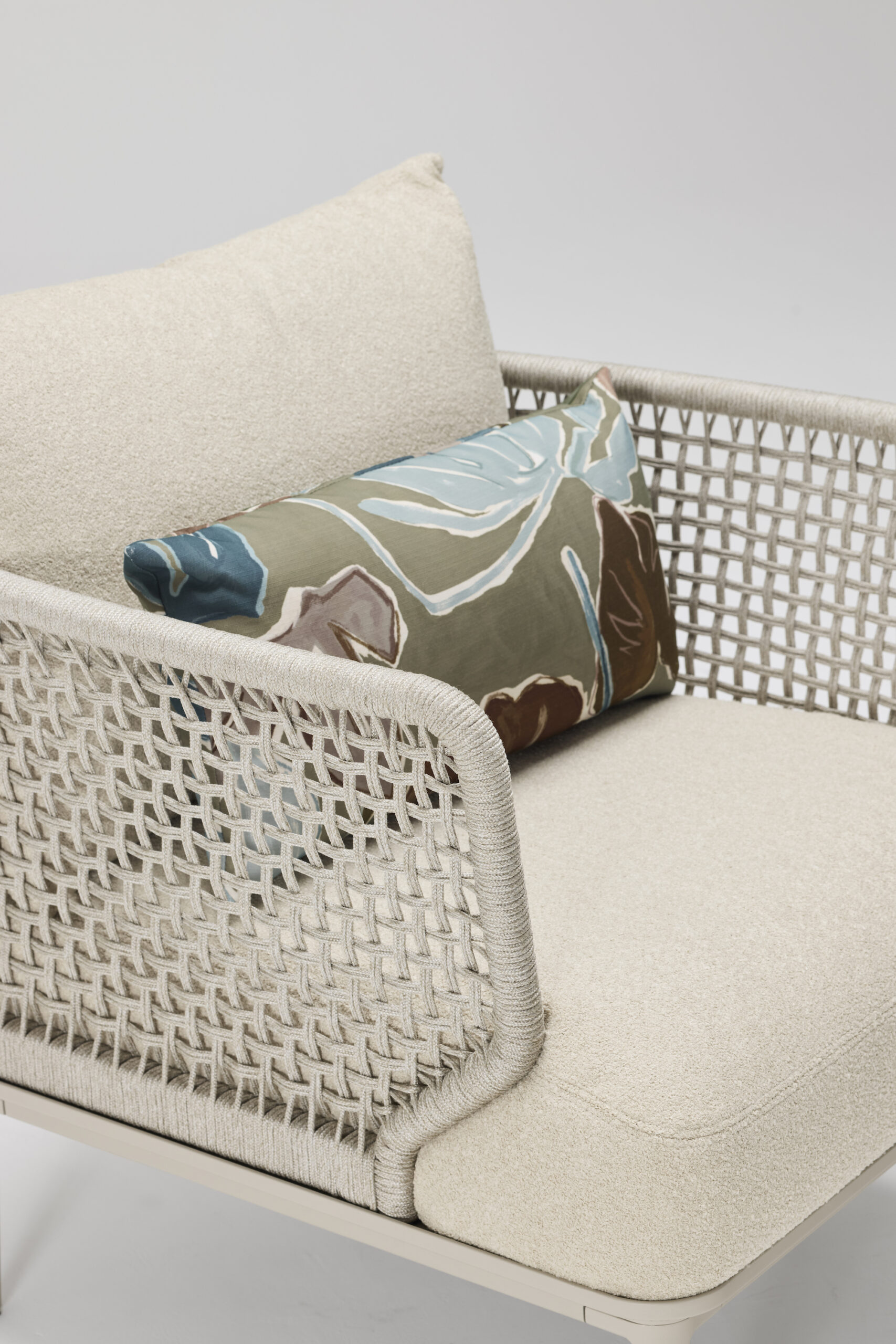 A modern beige armchair with woven sides, a large cream cushion, and a smaller decorative pillow featuring an abstract pattern in blue, green, and brown tones. The background is plain and light colored.