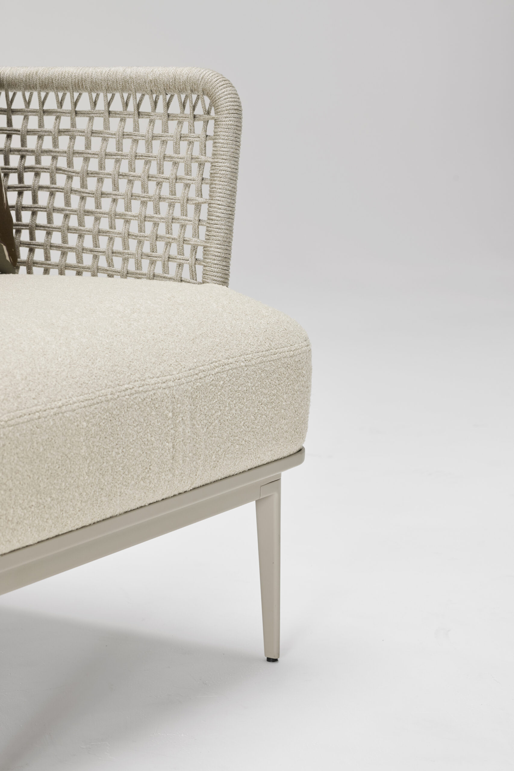 A close-up of a modern beige chair with a textured woven backrest and cushioned seat, set against a plain white background. Only part of the chair is visible, showing its sleek metal leg and contemporary design.