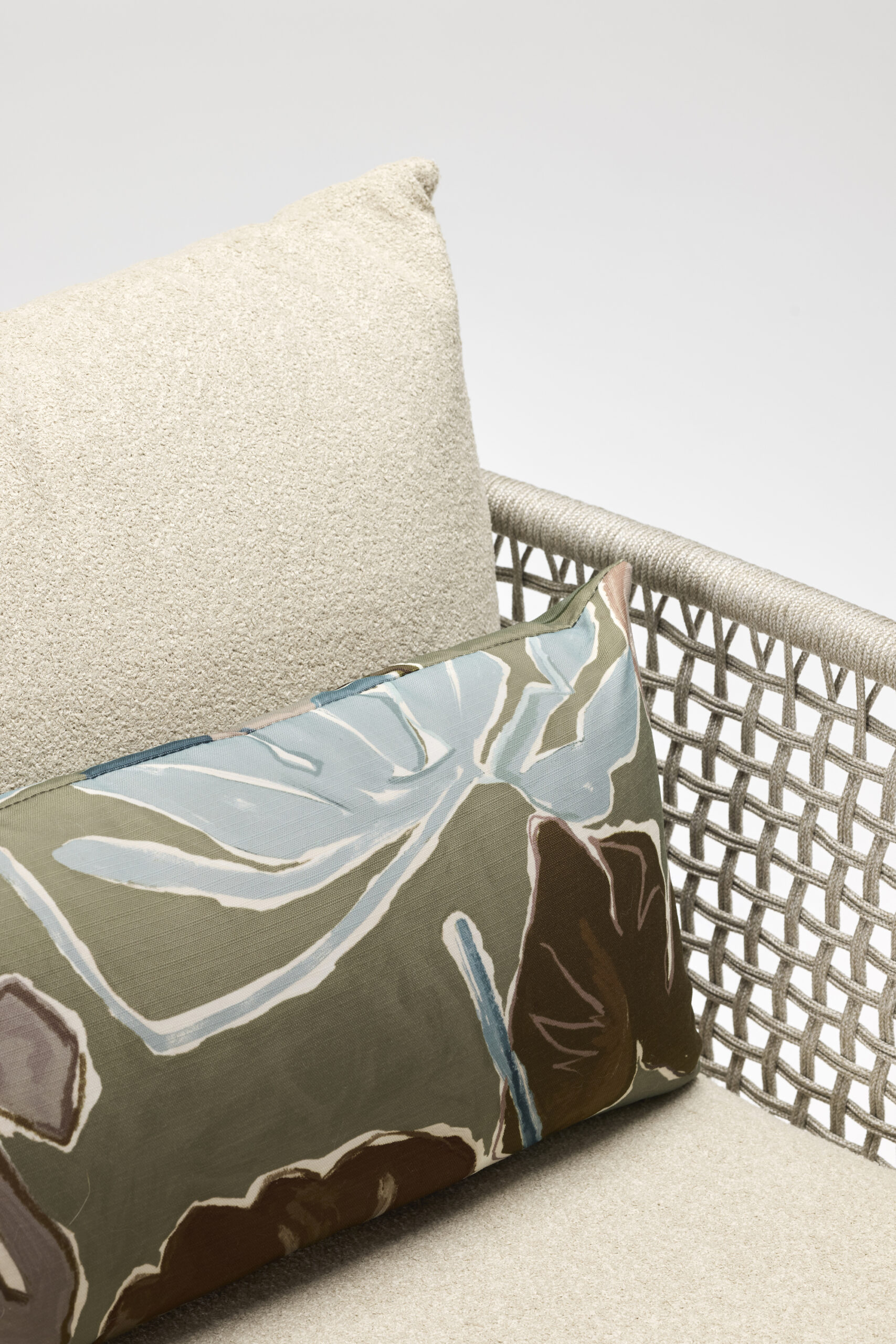 A beige cushioned chair with a woven rope armrest and a rectangular pillow featuring an abstract leaf pattern in green, blue, and brown tones. The background is plain and light.