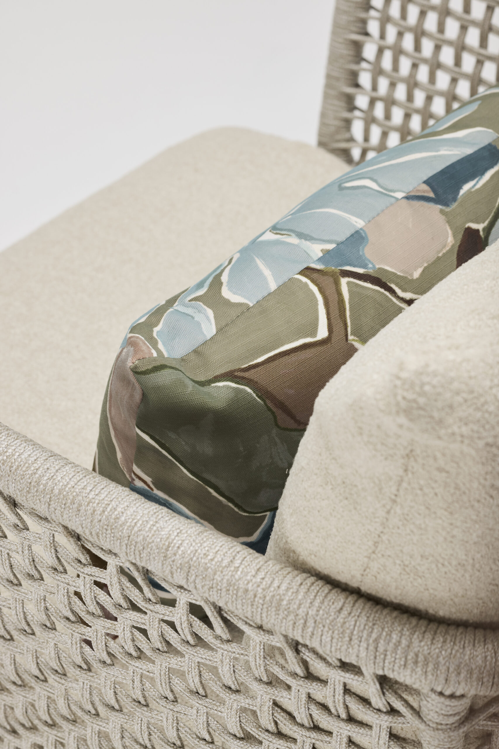 Close-up of a beige woven chair with a textured cushion and a decorative pillow featuring a green, blue, brown, and cream leaf pattern. The background is plain and white.