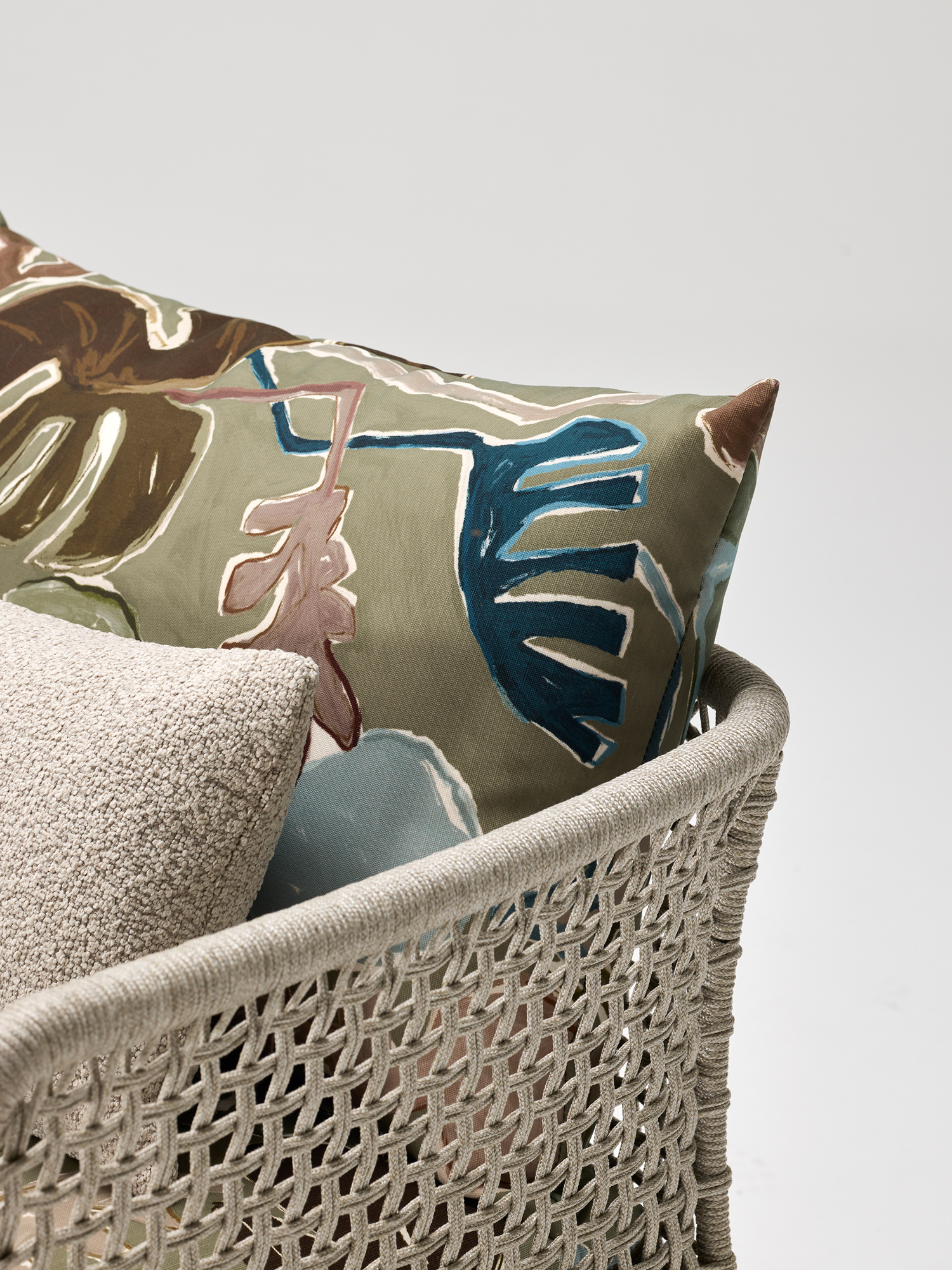 A close-up of a woven chair with a beige textured cushion and a decorative pillow featuring abstract, colorful leaf designs on a muted background, set against a plain light backdrop.