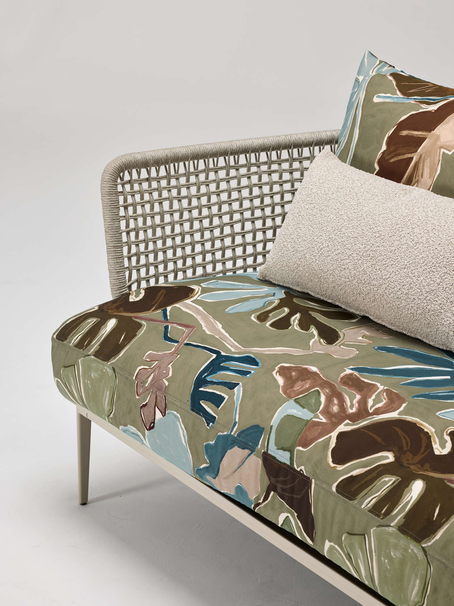 A modern lounge chair with a woven backrest, featuring a seat cushion and pillow covered in a leafy, abstract-patterned fabric in green, brown, and blue tones. A beige textured cushion rests against the pillow.