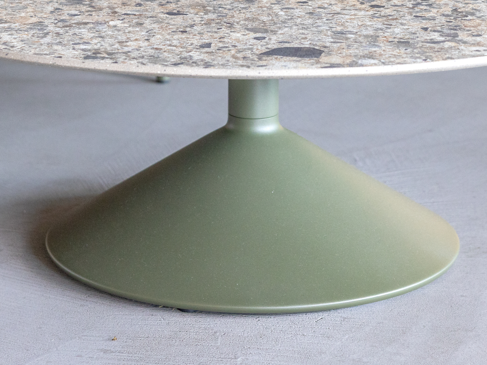 A close-up of a round table with a green, cone-shaped metal base and a grey speckled stone tabletop, set on a smooth, light-colored floor.
