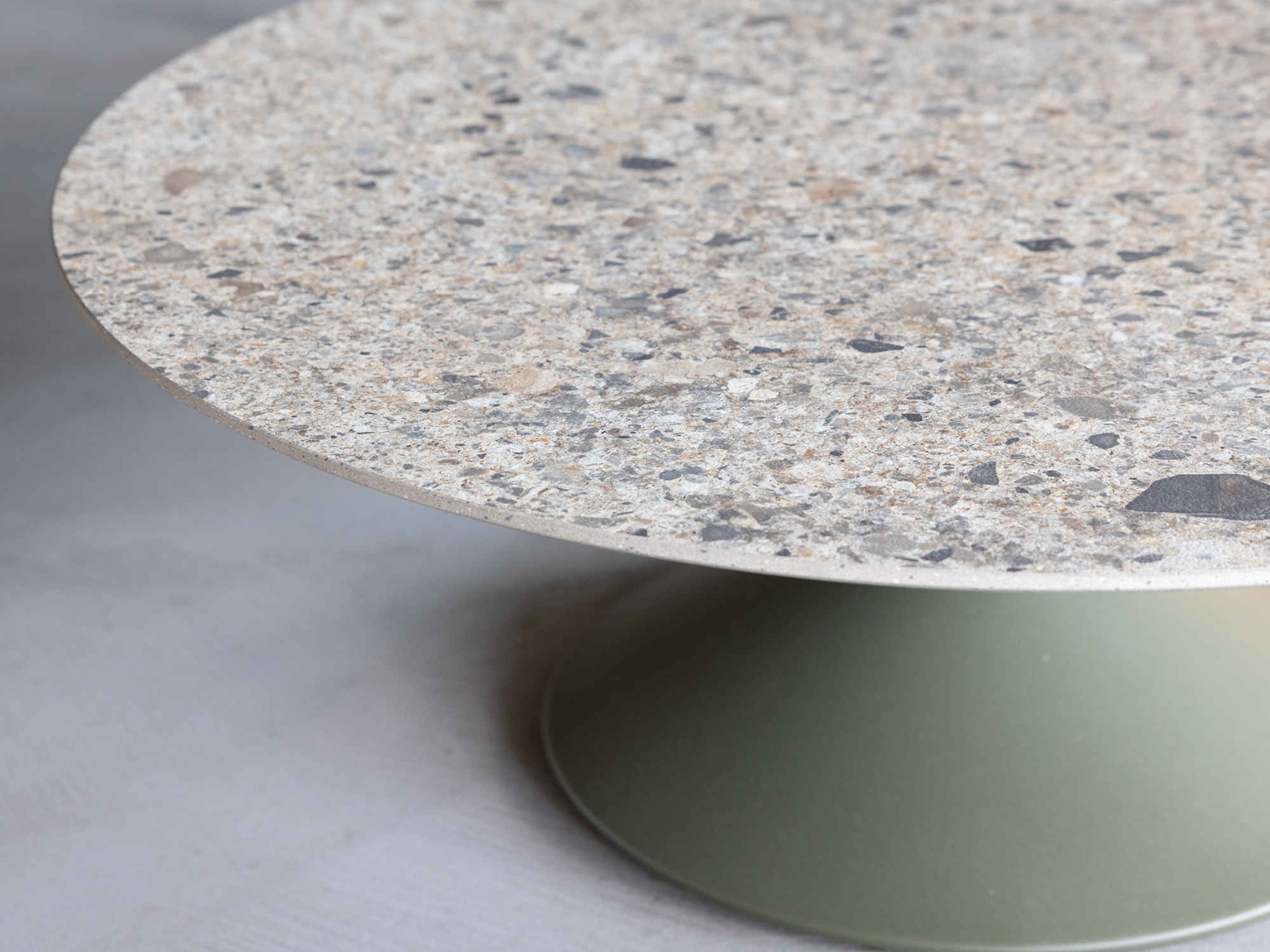A round table with a terrazzo-style stone top and a wide, matte green pedestal base, set against a light gray background.