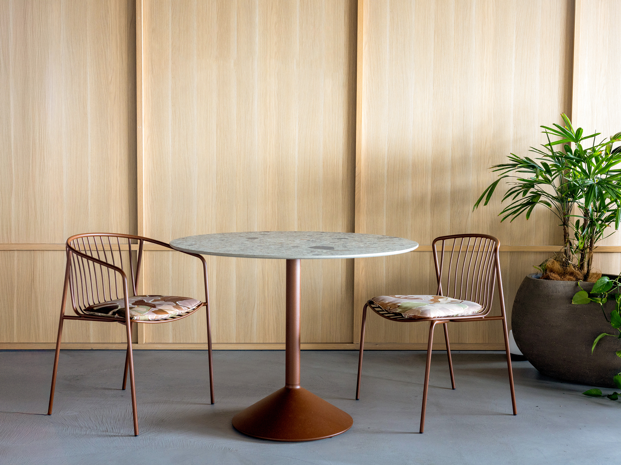 A round table with a marble top and two metal chairs with patterned cushions are set on a gray floor. A large potted plant is placed to the right, against a light wood-paneled wall.